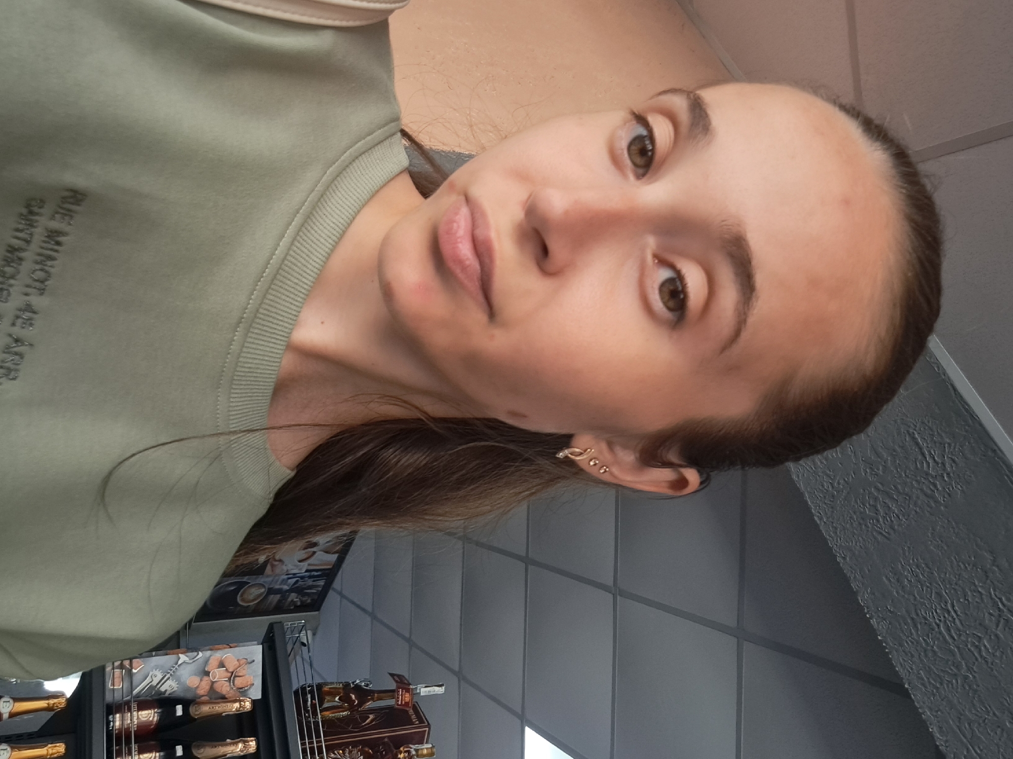 The image shows a medium shot of a young woman with long brown hair, wearing a light green sweatshirt. She has brown eyes and is looking directly at the camera. Her left ear is visible, adorned with two small earrings. The background appears to be an indoor setting, possibly a store, with a tiled ceiling and shelves displaying bottles and other merchandise. The lighting suggests it is daytime. There is no indication of the city of Bilhorod-Dnistrovskyi, Ukraine.