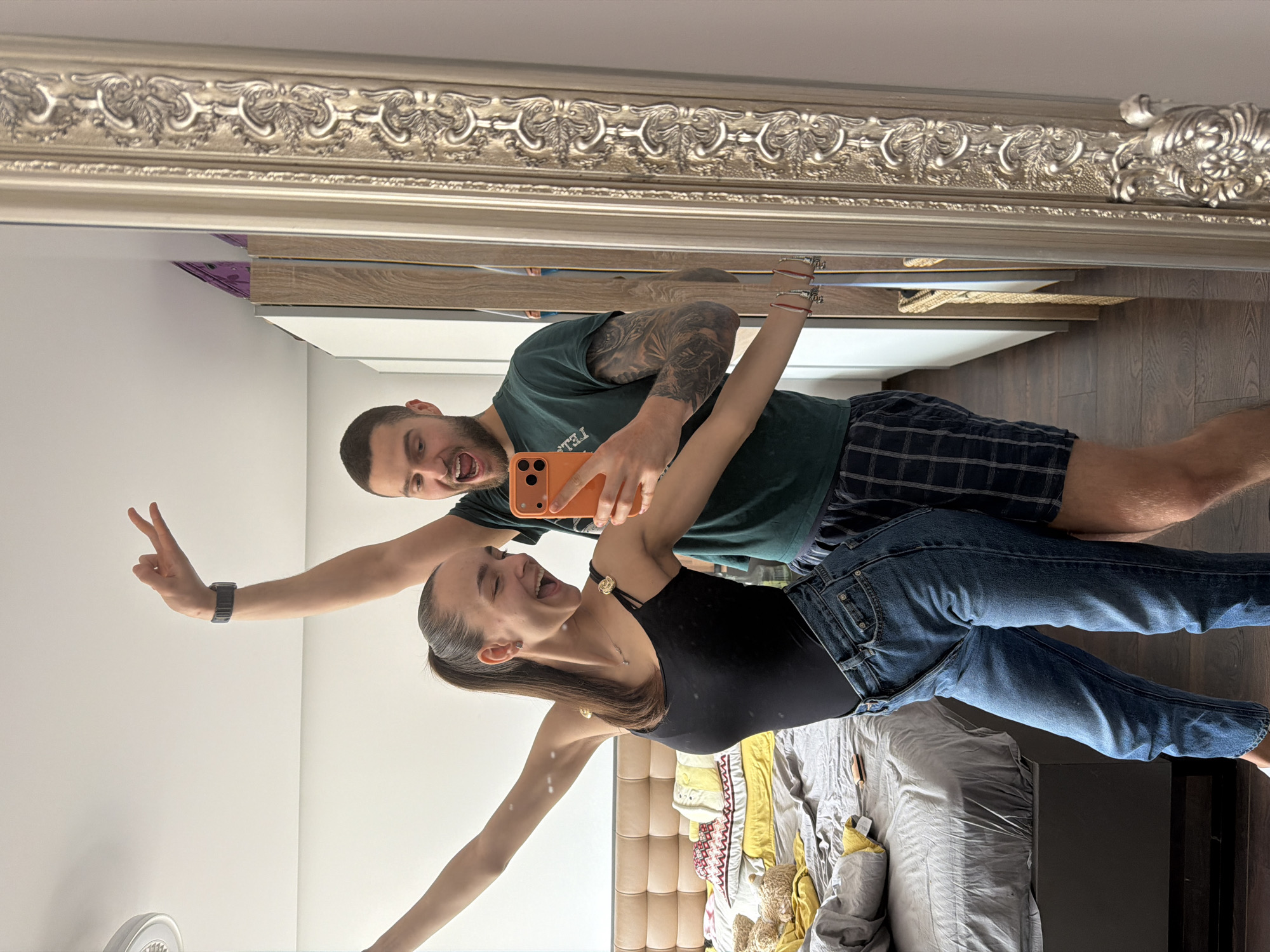 A couple poses for a mirror selfie in what appears to be a bedroom in an apartment or hotel room. The woman is wearing a black bodysuit and blue jeans, while the man is wearing a dark green t-shirt and plaid shorts. The woman is holding one arm up making a peace sign, and the man is holding the smartphone for the photo. The mirror is ornate with a silver frame. The bedroom has a bed with a gray comforter and yellow accents. A light-colored padded headboard is visible. There are light-colored cabinets above a shelf with wood elements. The location appears to be indoors, with a modern interior. It is not possible to determine the city of Sofia, Bulgaria from this image alone.
