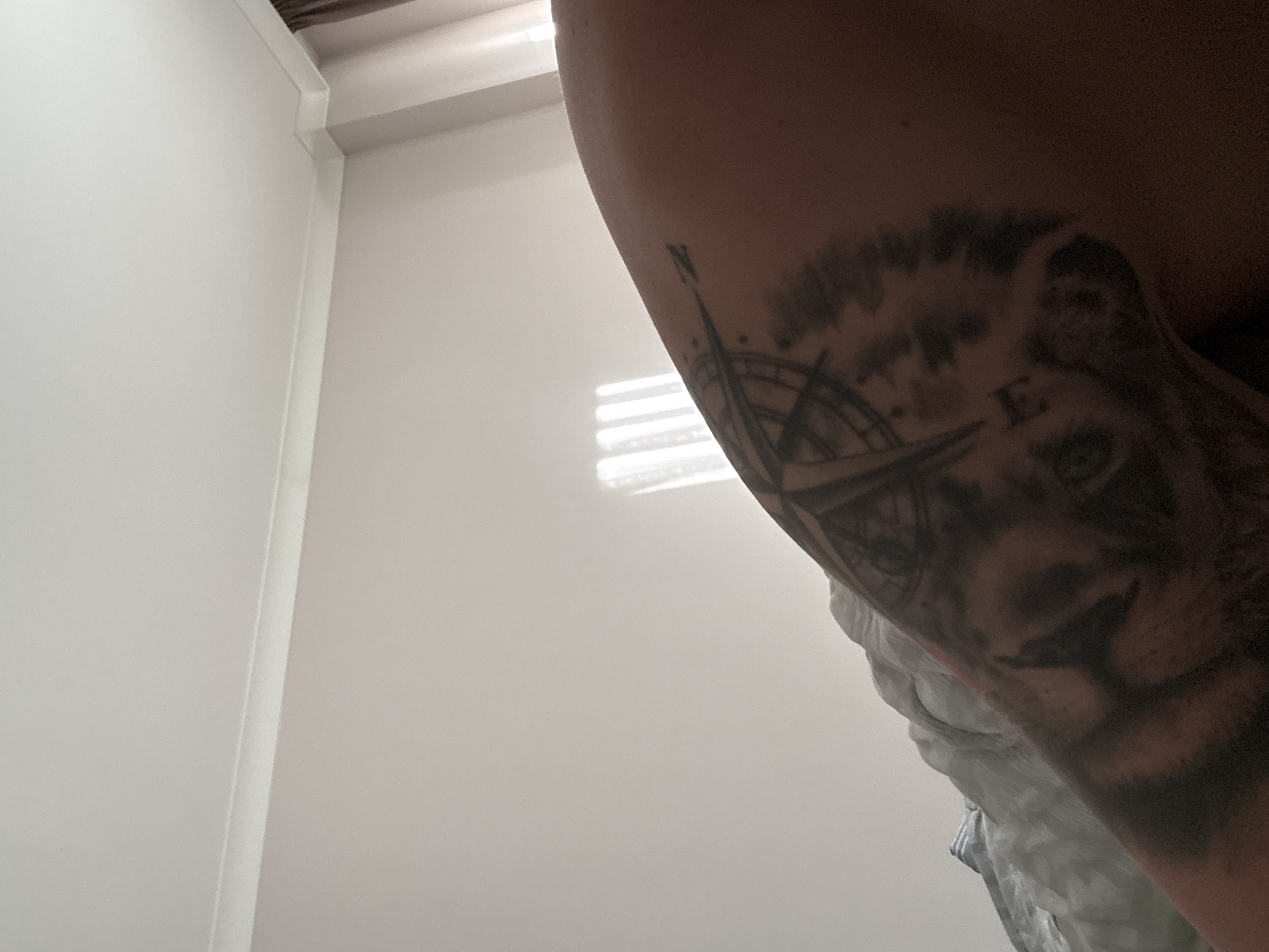 The image shows a close-up of an arm with a tattoo. The tattoo features a compass and the face of a lion. The background includes a section of a white wall and a window with slats. There is no information in the image indicating that this is in Varna, Bulgaria.
