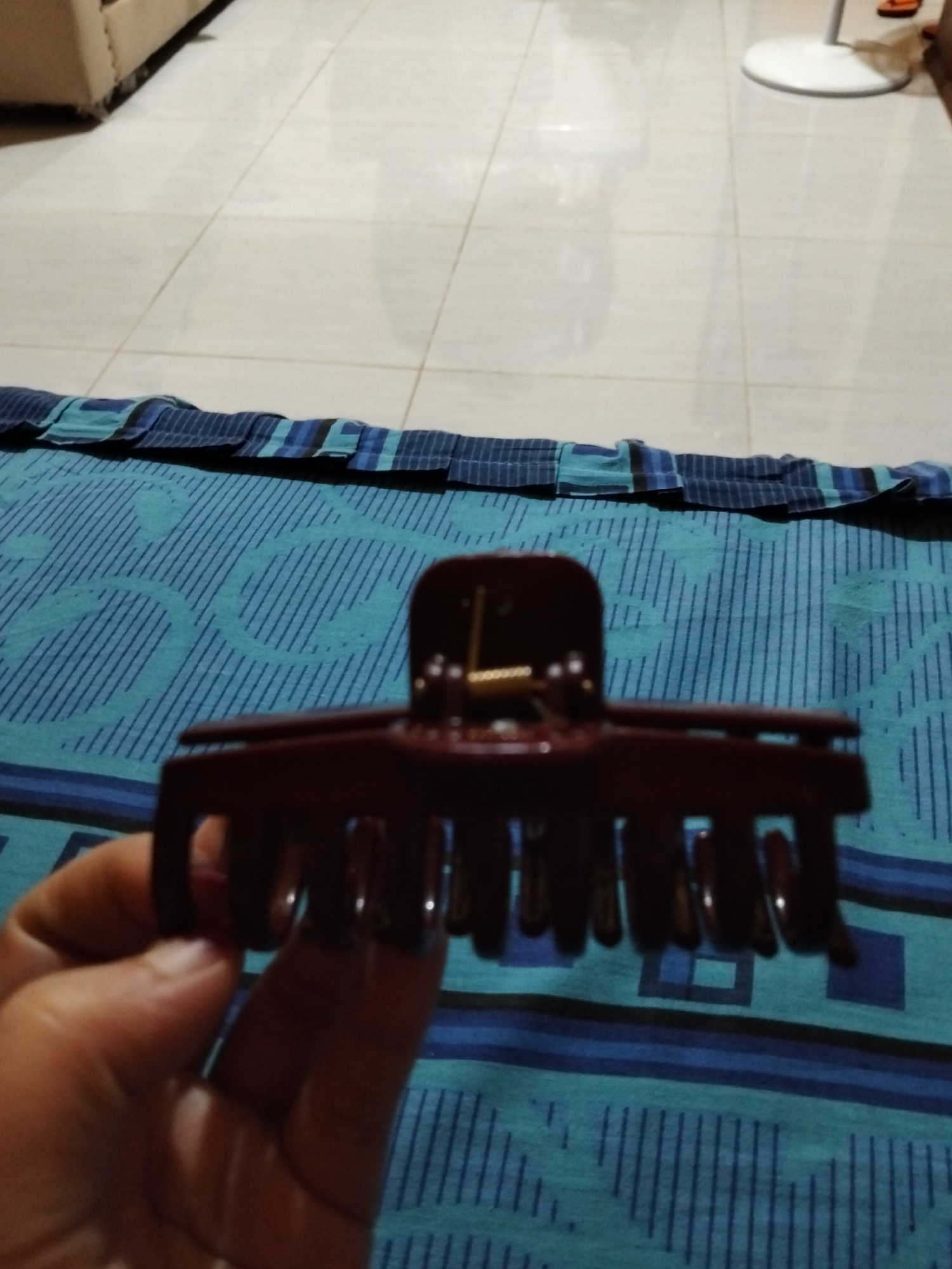 A hand is holding a maroon hair claw clip over a blue and teal patterned fabric, possibly a sarong or bedsheet, on a tiled floor. The hair clip is open and its teeth are visible. In the background, a portion of a beige sofa and a white floor fan are visible. The scene appears to be indoors, likely in a residential setting in Payagaboh, Indonesia.