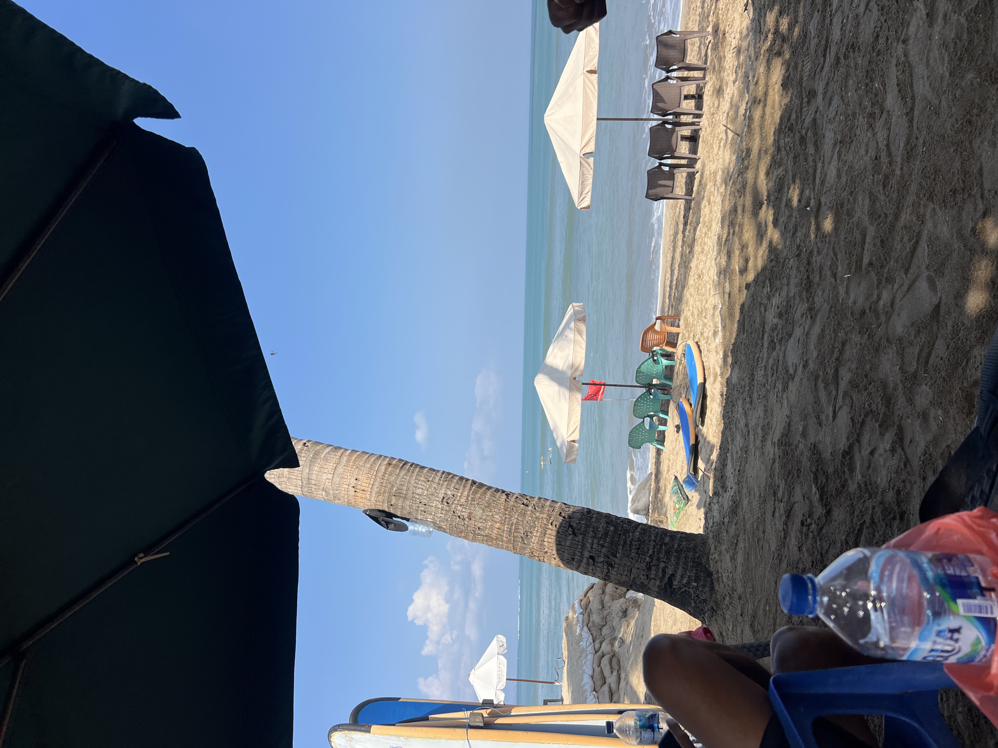 The image captures a serene beach scene in Kuta, Indonesia. The foreground is dominated by the sandy shore, with shadows cast from an overhead umbrella. Two individuals are partially visible, one with legs crossed in a blue plastic chair, and another with a hand and arm in the frame. A bottle of AQUA brand water is prominently placed in the foreground.
Beyond the immediate seating area, the beach stretches towards the ocean. Several surfboards stand upright against a palm tree. Beach chairs, both green and brown plastic, are arranged near the water's edge, some accompanied by light-colored beach umbrellas. The ocean is a calm turquoise, with gentle waves lapping the shore. A few people can be seen swimming or wading in the distance. The sky is a clear, bright blue, with a few wispy clouds. Sandbags are visible near a white umbrella further down the beach. The overall atmosphere is one of relaxation and tropical leisure.