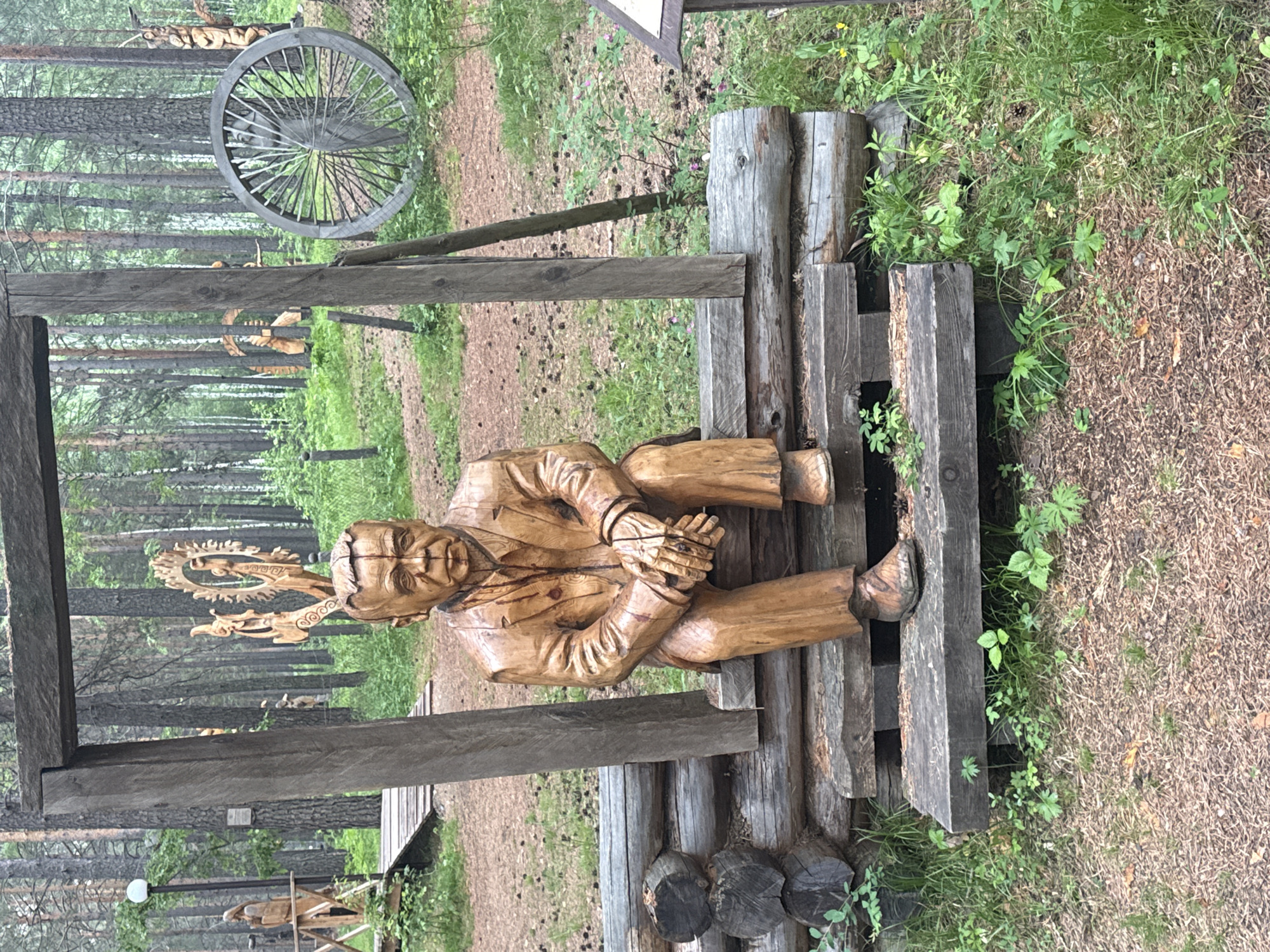 The image shows a wooden sculpture of a seated man in a forest setting, likely in the city of Savvateevka, Russia. The sculpture depicts a man with a serious expression, wearing a suit and tie, with his hands clasped in his lap. He is seated on a bench made of logs. Behind the sculpture, a wooden archway frames the scene. In the background, there are more wooden sculptures, including a large spoked wheel and other figures, set amongst tall pine trees. The ground is covered with pine needles and grass. The overall atmosphere is rustic and artistic.
