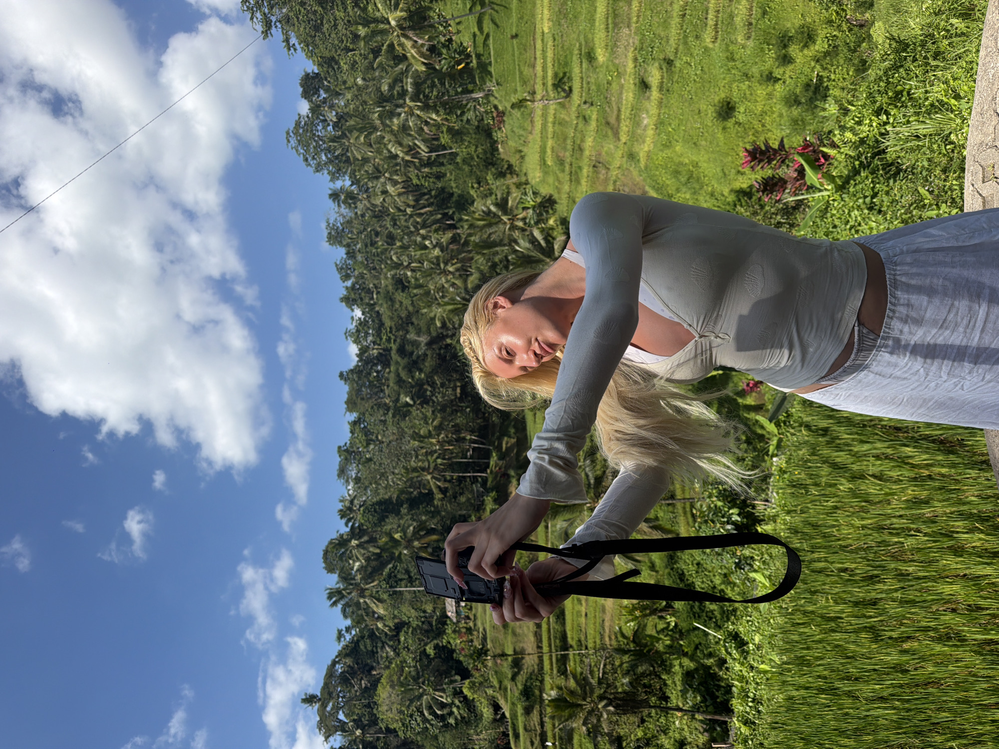 A blonde woman is taking a selfie with a camera in the Tegallalang rice terraces in Indonesia. The woman is wearing a white long-sleeved shirt and a white skirt. She is standing in front of a lush green landscape of rice paddies and palm trees. The sky is blue with scattered clouds. The image is taken from a low angle, looking up at the woman and the sky.