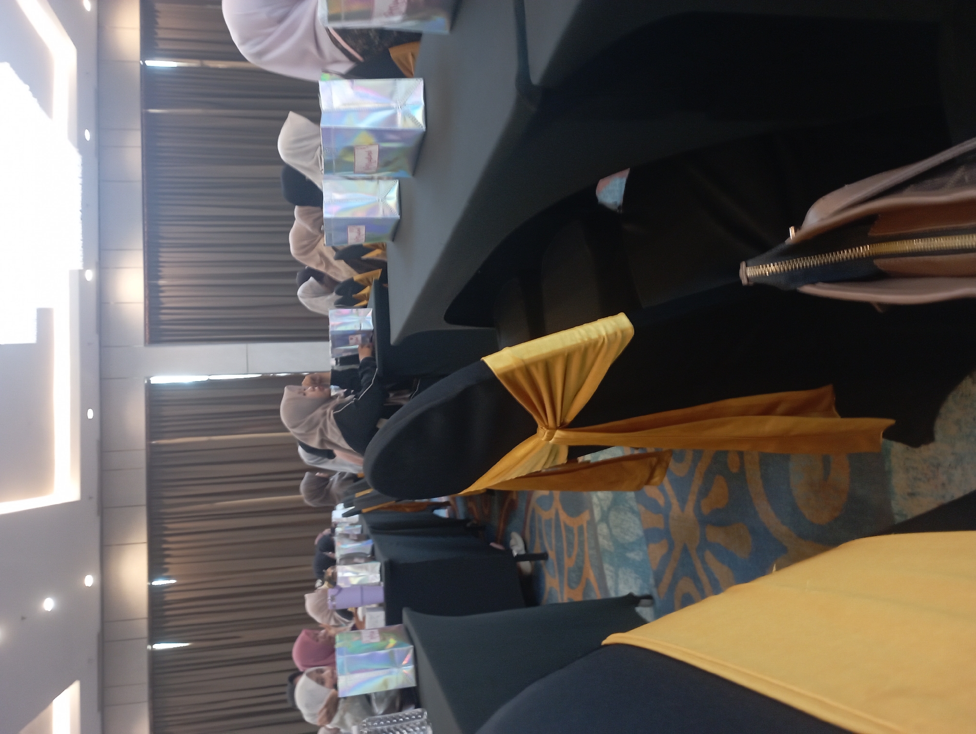 The image depicts a dimly lit indoor event space, likely a conference room or banquet hall, in Purwakarta, Indonesia. Several women, predominantly dressed in hijabs, are seated at tables covered with black tablecloths. The tables are adorned with holographic gift bags. Chairs are draped with yellow sashes. The room is furnished with a patterned carpet and features large curtains in the background, suggesting a formal setting. The lighting appears to be artificial, coming from overhead fixtures. A purse with a gold zipper is visible in the foreground.