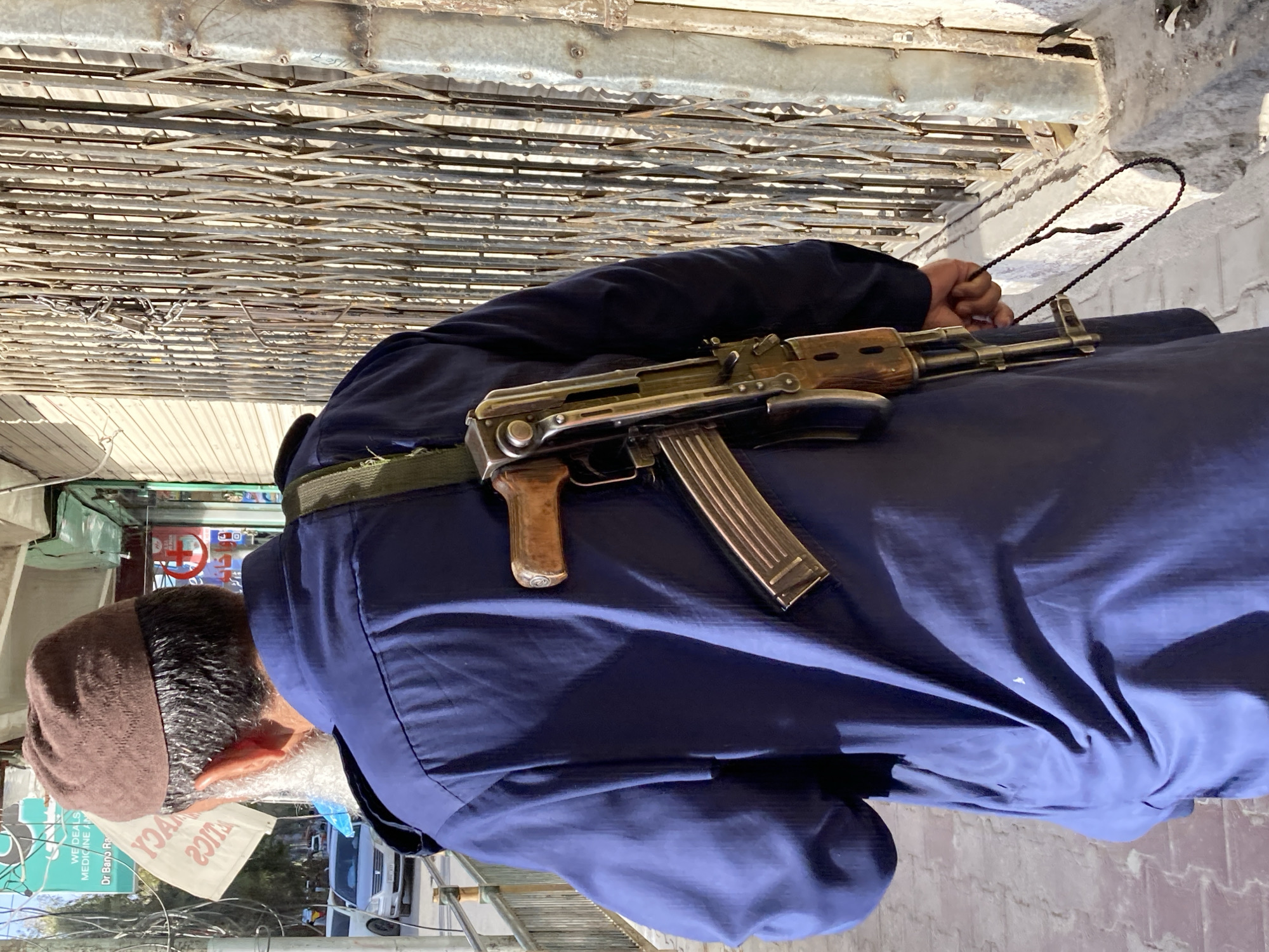 A man wearing a brown knit cap and a blue overcoat is walking in what appears to be a city alleyway. He is carrying a rifle strapped over his shoulder. In the background there is a pharmacy, and metal roller doors above and to the side. Based on the pharmacy signage, the location is Quetta City Tehsil, Pakistan.