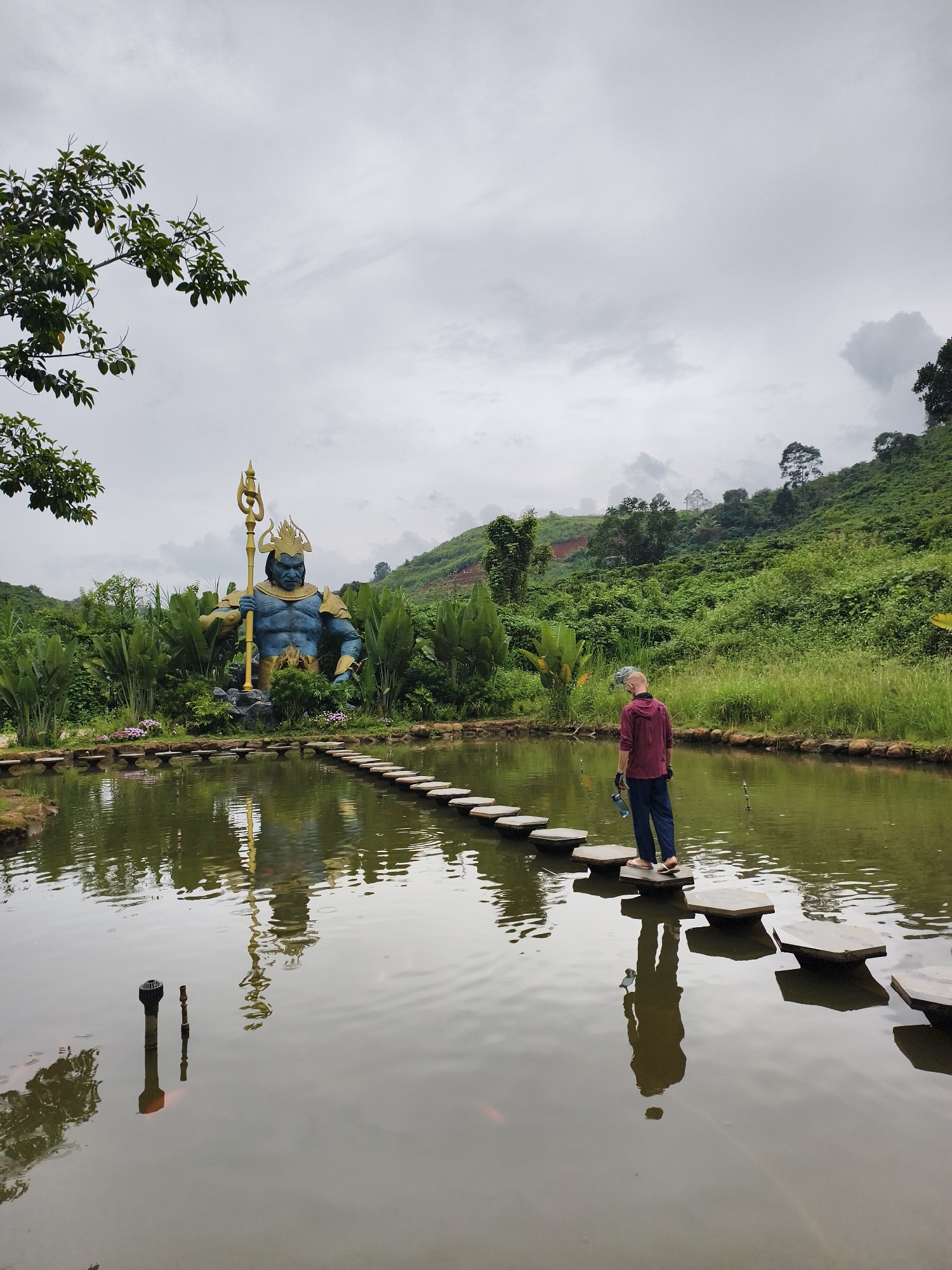 A person walks across stepping stones in a pond toward a large blue statue in Thôn 1, Vietnam. The statue, depicted as a muscular figure, holds a golden trident. Lush greenery and a hillside serve as the backdrop. The pond's surface reflects the statue and the walking person. A cloudy sky is visible overhead.