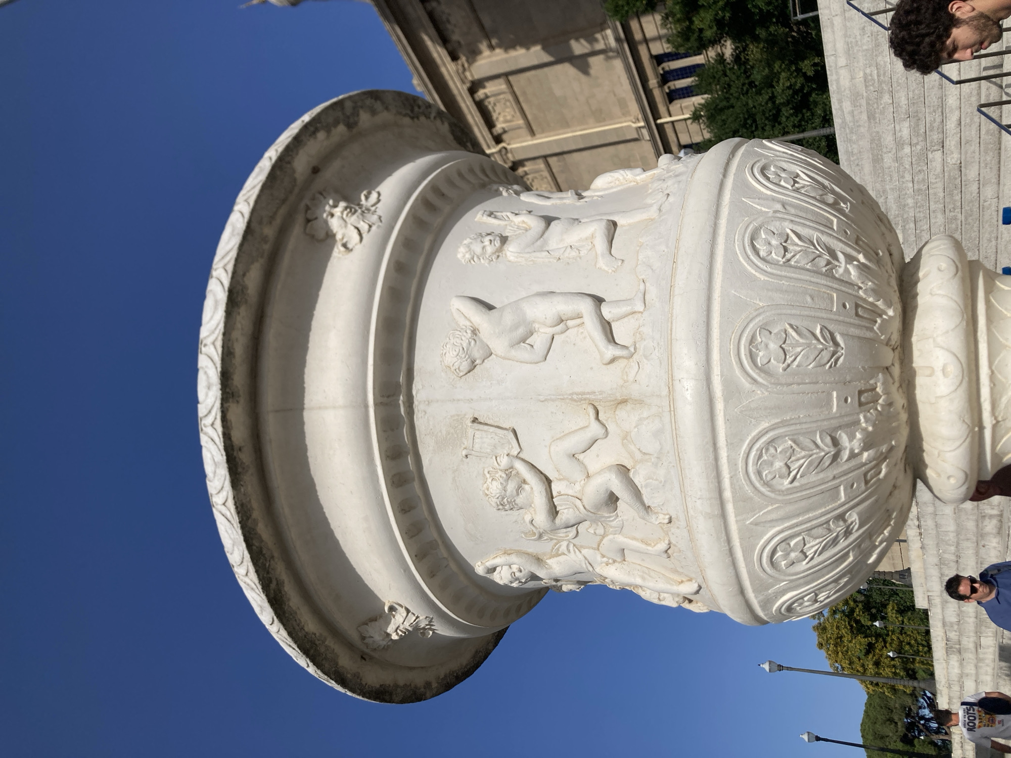 A large, white, ornate urn with bas-relief carvings of cherubic figures is the central focus of the image. The urn is positioned in the foreground, with its detailed carvings, including a cherub playing a lyre, clearly visible against a bright blue sky. The urn is part of the exterior of a large, classical-style building, likely the Palau Nacional, located in Barcelona, Spain, on Montjuïc hill. Behind the urn, a wide set of stone stairs ascends, flanked by lampposts. In the mid-ground, several people are visible walking up the stairs. To the right of the urn, a man with dark curly hair is partially visible in profile, looking towards the urn. The lighting suggests it is daytime.