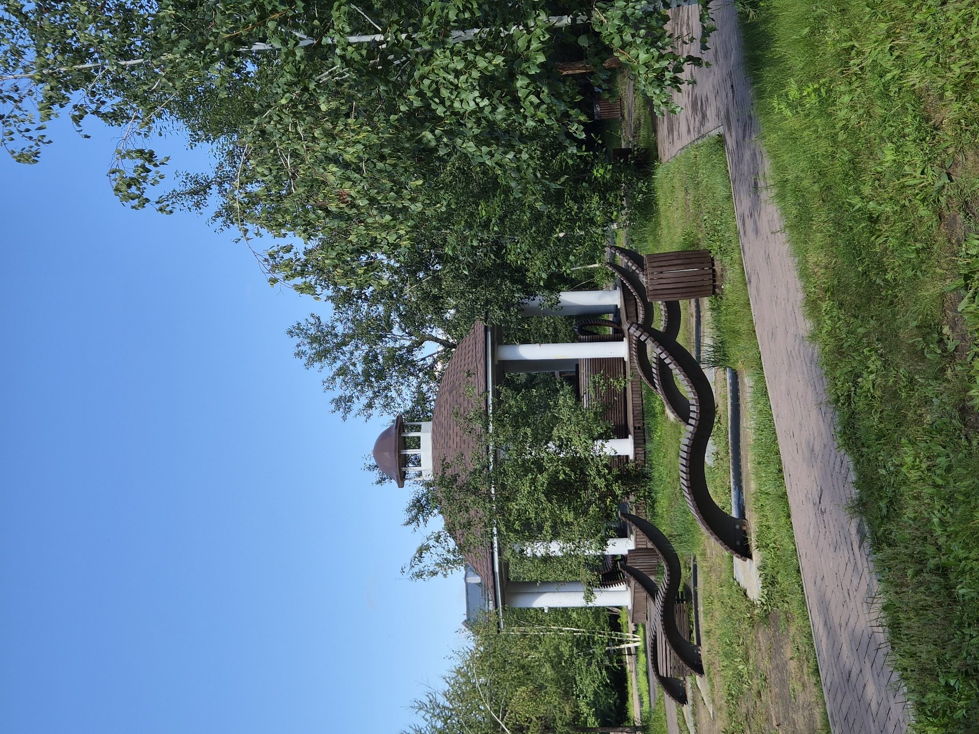 The image depicts a park scene on a clear day. In the center is a white gazebo with a brown, octagonal roof, surrounded by lush green trees. In front of the gazebo, there are several curved, wooden lounge chairs placed on concrete pads. A brick pathway winds through the grassy area, and a wooden trash bin is visible to the right of the pathway. The sky is a vibrant blue with no clouds. The overall impression is one of a well-maintained, tranquil public space. This park is located in Chita, Russia.