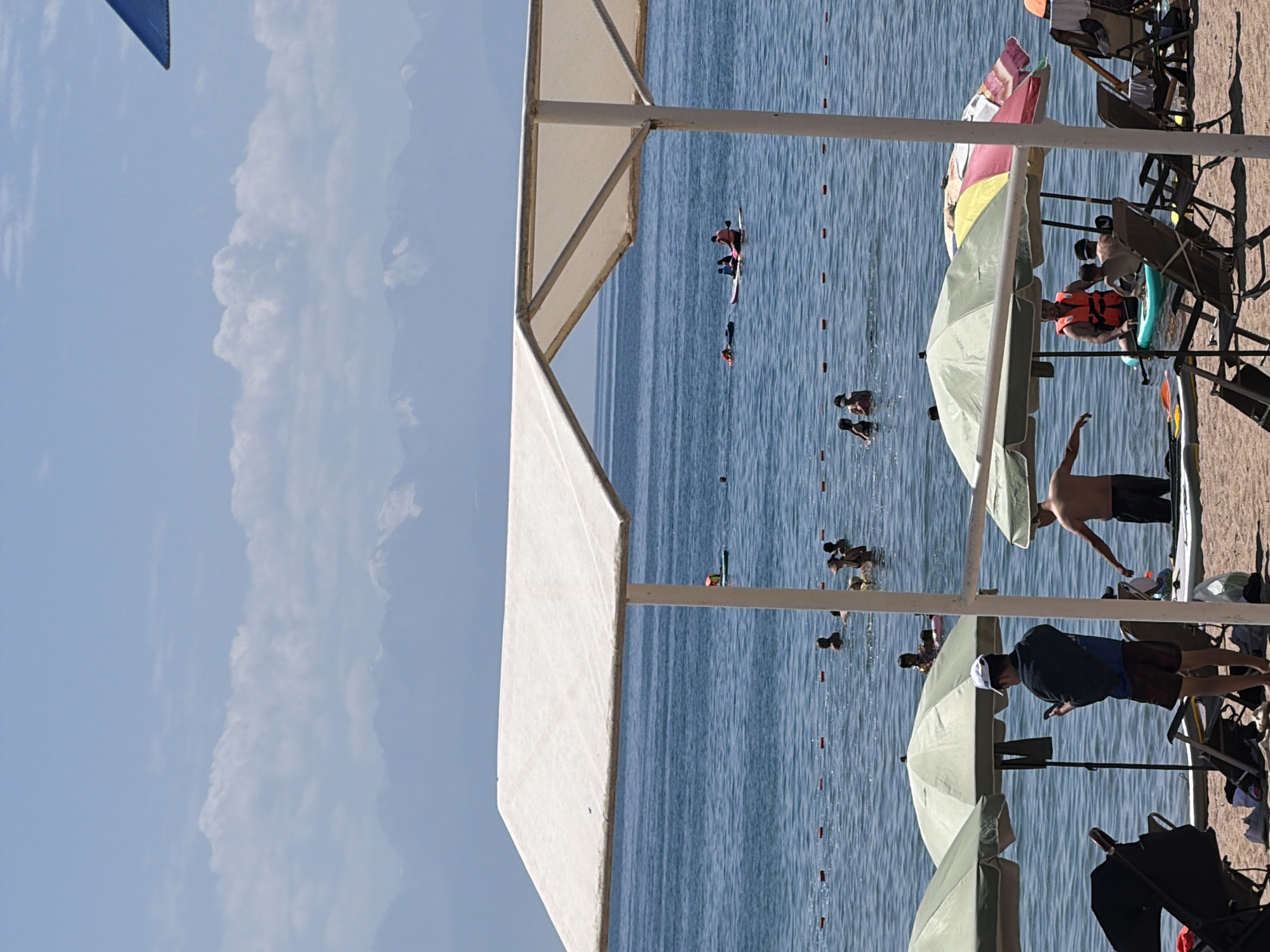 In the city of Unknown, Unknown, a beach scene unfolds under a bright, clear sky with distant snow-capped mountains and scattered clouds. The foreground is dominated by a large, white canopy supported by poles, casting a shadow over the sandy beach. Umbrellas, primarily green and one with colorful stripes, are set up on the sand, with lounge chairs visible underneath some.
The calm blue water of a lake or sea stretches out to the horizon. Several people are enjoying the water. Some are swimming, while others are engaged in water activities. A person is visible on a paddleboard, and another group is on a larger paddleboard or small boat. Red buoys mark a boundary in the water.
On the beach, several individuals are present. One man wearing a white cap and holding a phone stands near a green umbrella, facing the water. Another man, shirtless, stands in the water near a paddleboard. A baby stroller is parked on the sand, and other beach chairs are occupied or empty. The overall scene depicts a leisurely day at the beach with various individuals and groups enjoying the water and sun.