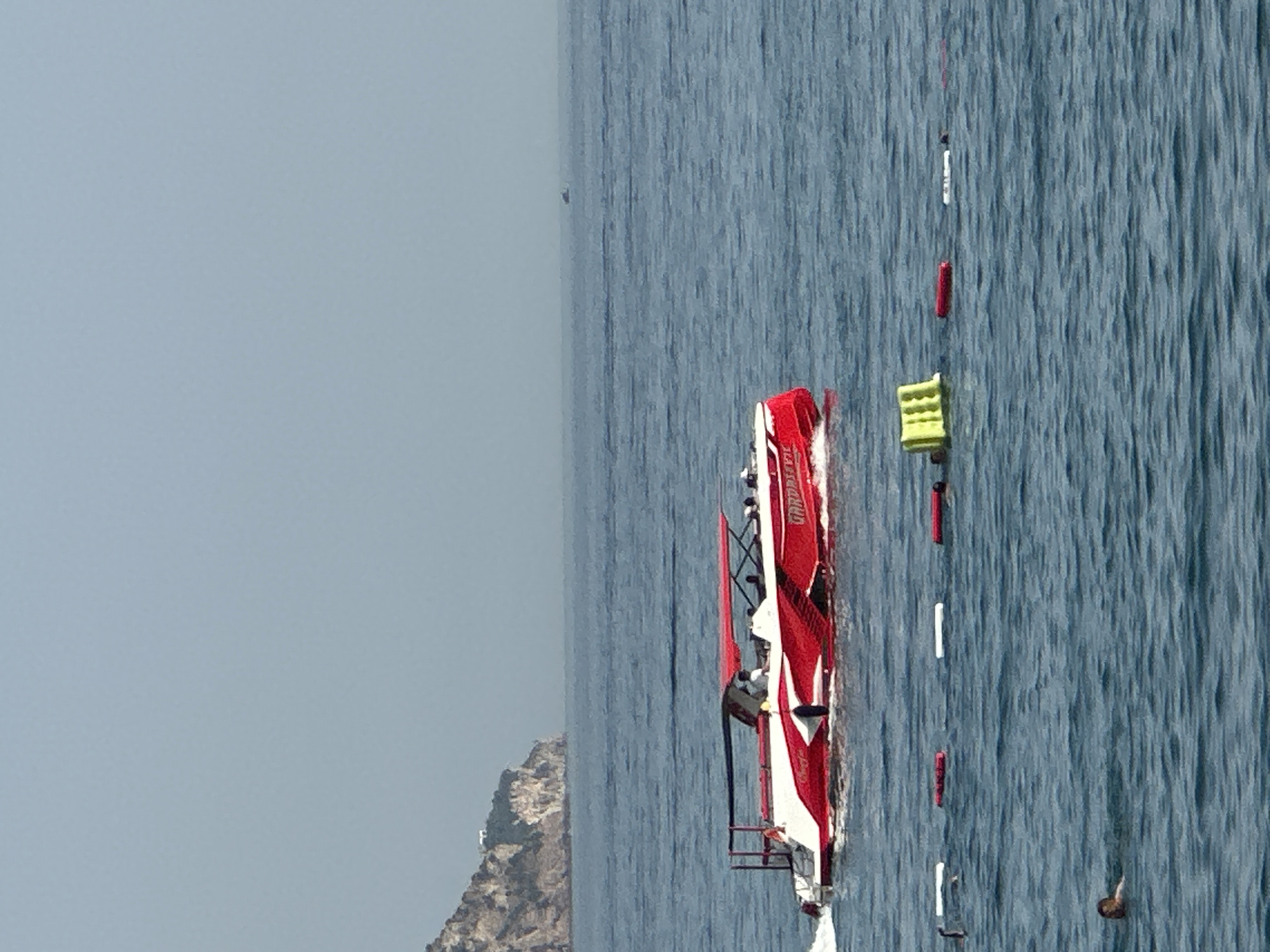 The image depicts a vibrant red and white speedboat navigating calm, blue waters near a rocky coastline, likely in Boreti, Montenegro. The boat, a racing model with a sleek design, is in motion, creating a visible wake. Several people are visible on board, appearing to be enjoying a boat trip or participating in a water sport. A series of buoys, some red and some white, are spaced out in the water, possibly marking a course or a specific area. A large, rugged rock formation rises from the water on the left side of the frame, providing a sense of the coastal environment. The sky is a hazy, light blue, suggesting either a slightly overcast day or a distant haze, possibly indicating it is mid-day. The overall impression is one of leisure and activity on the water, with the bright colors of the boat and buoys contrasting with the natural elements of the sea and coastline. No discernible text is visible on the boat or other elements in the image.