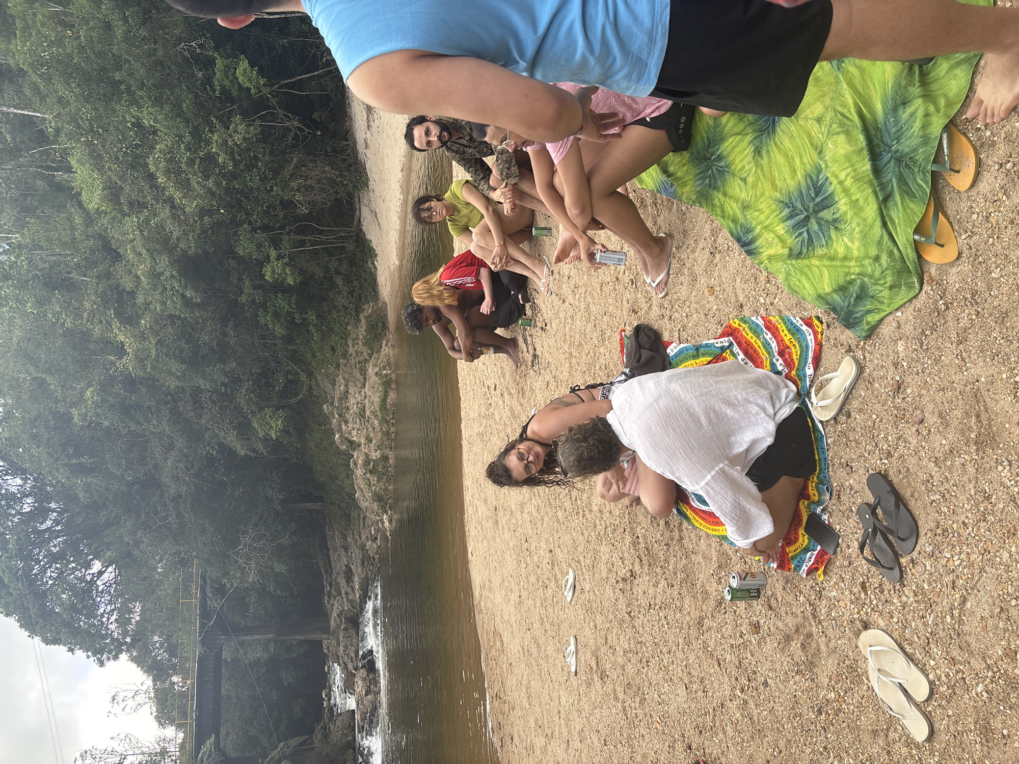 A group of people are gathered on a sandy bank next to a body of water, likely a river or lake, with trees visible in the background. From foreground to background, on the right side of the frame, are: a woman lying down with her head looking at the camera; a person in a white shirt crouched near a colorful blanket; a pile of flip-flops; a group of people sitting together on a green patterned cloth or blanket. Two or three beer cans are lying on the ground. The water appears to have a slight brownish tint. A bridge is slightly visible on the left.
The location may be Parque Estadual da Cantareira, São Paulo, Brazil.