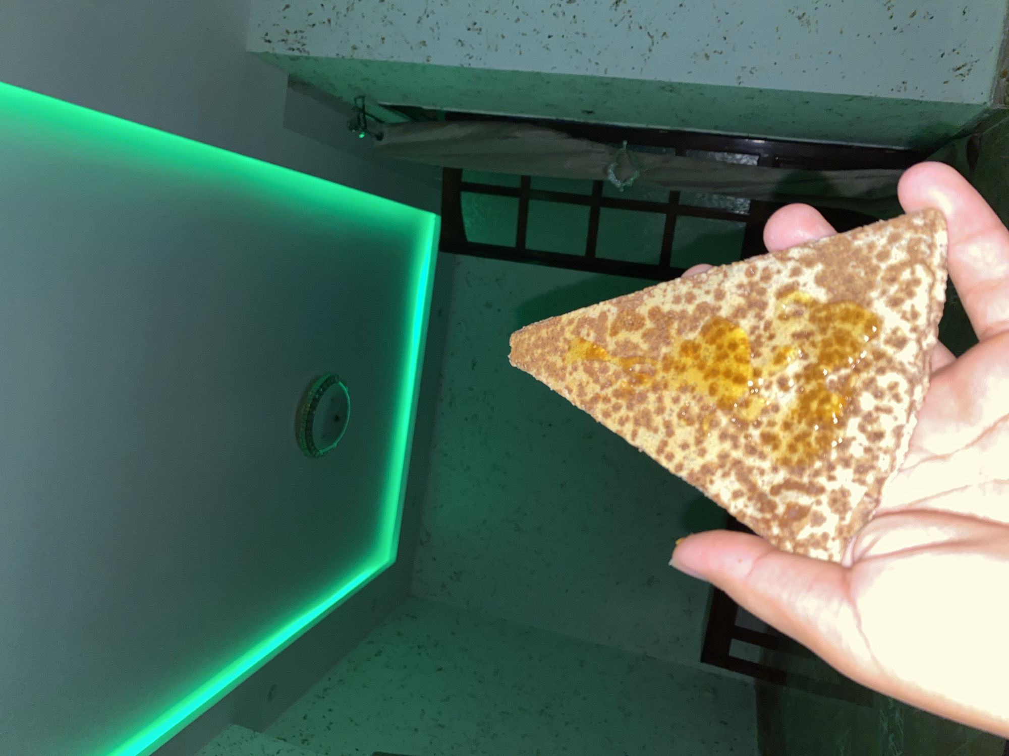 A hand is holding a triangular piece of food, possibly a pastry or flatbread, topped with a golden syrup and speckled with brown crumbs. The background features a room illuminated by green LED strip lighting along the ceiling. A ceiling light fixture is visible, along with a textured wall. To the right, a wooden door with glass panes is partially obscured by a light-colored curtain. In the background, a wooden chair or stool is also present. The scene appears to be indoors, likely in a residential setting within the city of Baraki, Algeria.