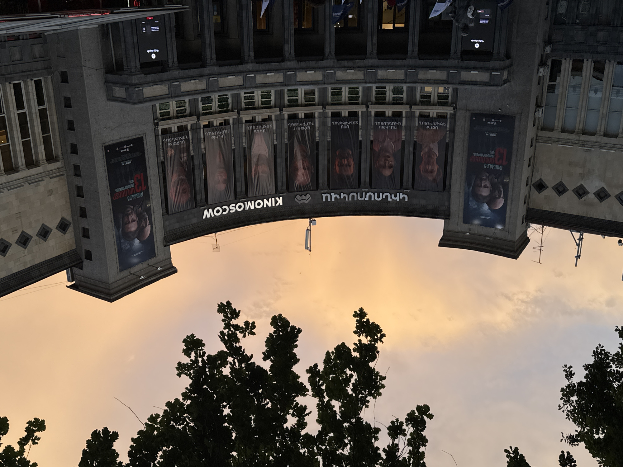 The image shows the "Moscow Cinema" in Yerevan, Armenia. The photo is taken from a low angle, looking up, and the image is rotated 180 degrees. The architecture of the cinema building is visible, including posters and banners displayed along the facade. In the foreground, there are tree branches silhouetted against the evening sky, which is tinged with shades of orange and grey. The cinema's name, "KINOMOSCOW", is displayed prominently. Banners feature portraits of various individuals and Armenian text. A poster on the left is related to a movie titled "13 Kilometers of deadly distance."