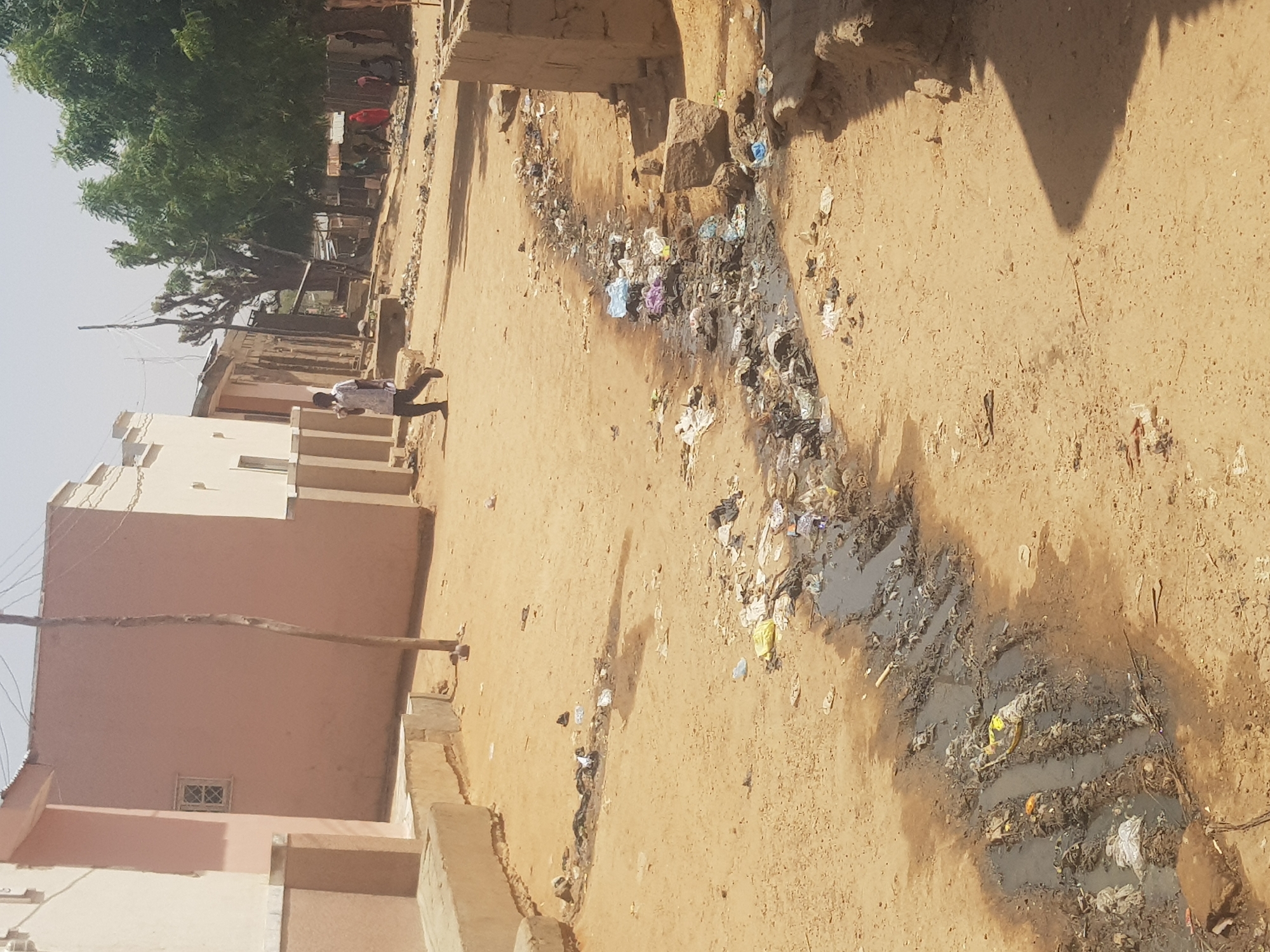The image shows a dirt street in Dutsinma B, Nigeria. Litter and debris are scattered along a channel running down the center of the street. Water accumulates in the channel, creating a muddy, unsanitary appearance. Buildings line the side of the street, with visible walls and roofs constructed from clay and concrete. A person walks along the street toward the background. A tree line is visible further in the background. The environment appears dry and dusty.