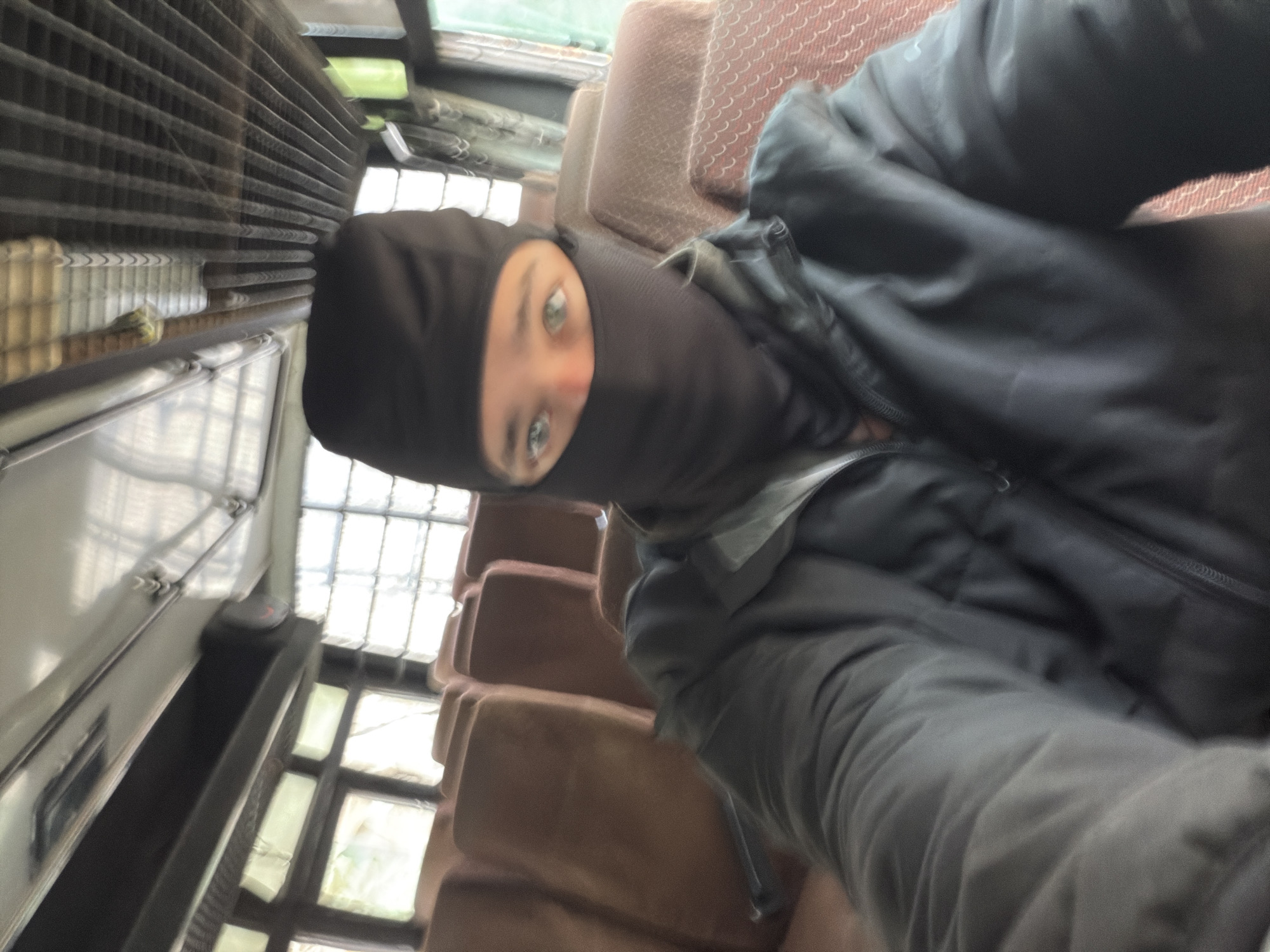 A young person wearing a black balaclava and a black jacket is seated on a bus. The person's eyes are visible through the balaclava opening. The bus interior features rows of brown seats with a patterned design. The windows appear to be along the side of the bus, and the ceiling has a ribbed metallic structure. The scene is captured from a first-person perspective, suggesting a selfie. The location is identified as Gayk, India.