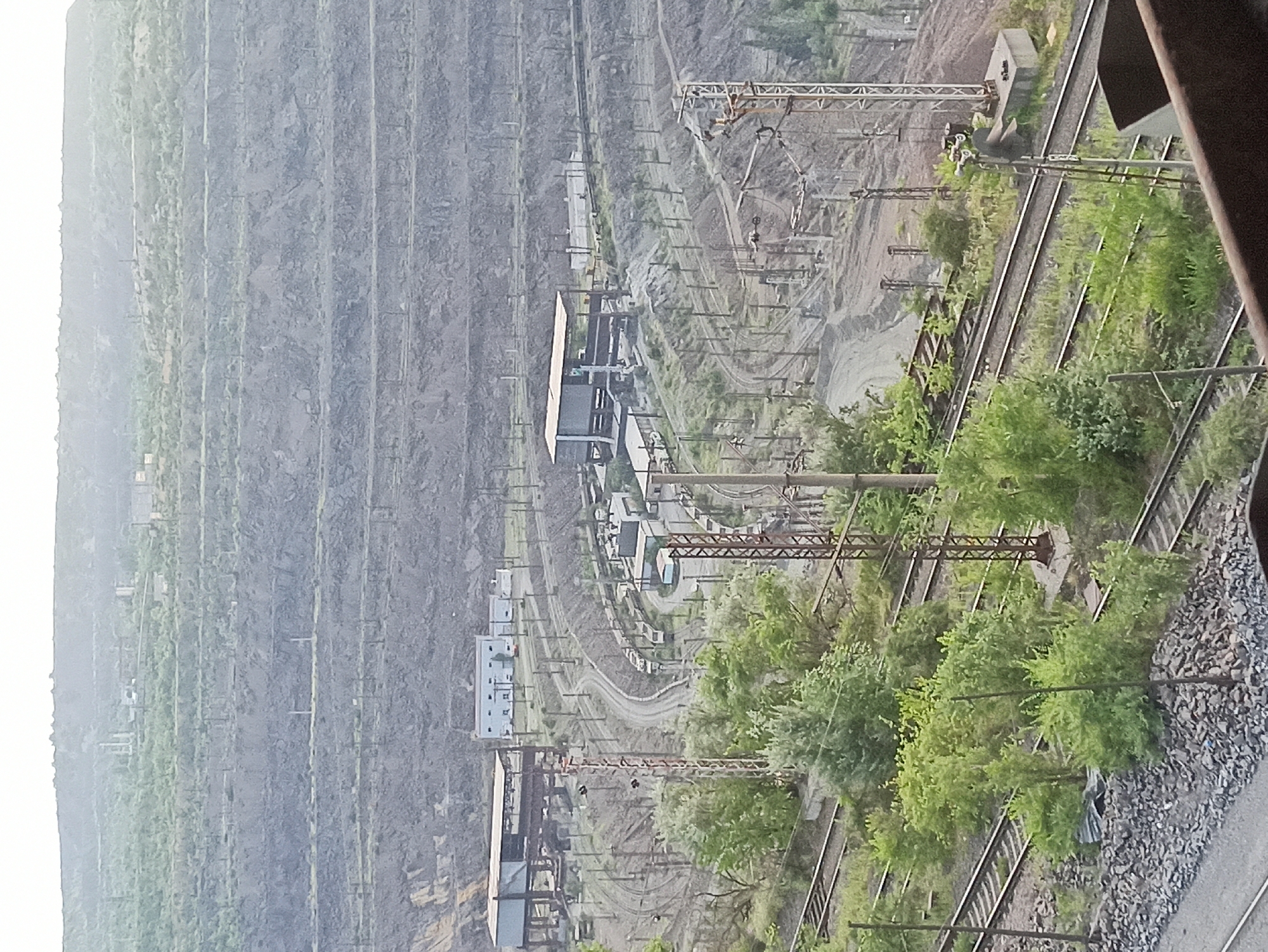This is a photograph of a mining operation or industrial site. The scene is dominated by vast, tiered excavations in a rocky hillside, indicative of large-scale resource extraction. Numerous sets of railroad tracks run through the site, suggesting transportation of materials. Several industrial buildings and structures, including what appear to be processing plants or storage facilities, are visible among the tracks and excavated areas. Overhead power lines and supporting poles are also prevalent, indicating ongoing industrial activity. Lush green vegetation, including trees and bushes, is growing on the slopes and around the industrial infrastructure, suggesting that the operation may be partially disused or that nature is reclaiming the area. The image is taken from an elevated perspective, looking down and across the site. The lighting suggests it is daytime, but the overall atmosphere is somewhat hazy or overcast. No people are clearly visible, and there is no discernible text in the image. The composition focuses on the scale and industrial nature of the landscape.