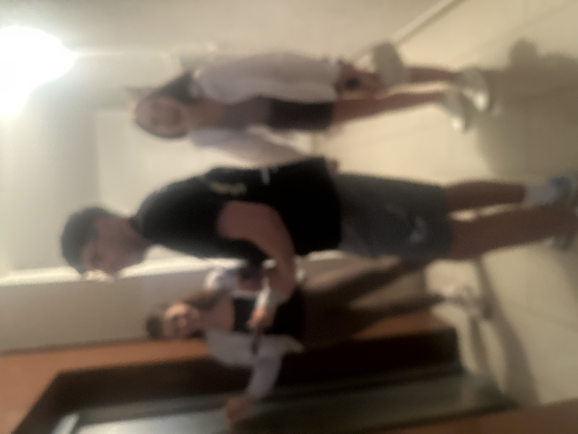 The image depicts three individuals in a hallway. A male is positioned in the foreground, wearing a dark t-shirt and grey shorts. To his left, a female is visible, wearing a light-colored jacket over a dark top and brown trousers. To the right of the male, another female is seen wearing a light-colored jacket over a dark top and dark shorts, carrying a small handbag. The hallway appears to be indoors, with a door on the left and a bright light source at the end of the corridor. The overall scene is out of focus. There is no information present in the image to indicate a location in the city of Shumen, Bulgaria.