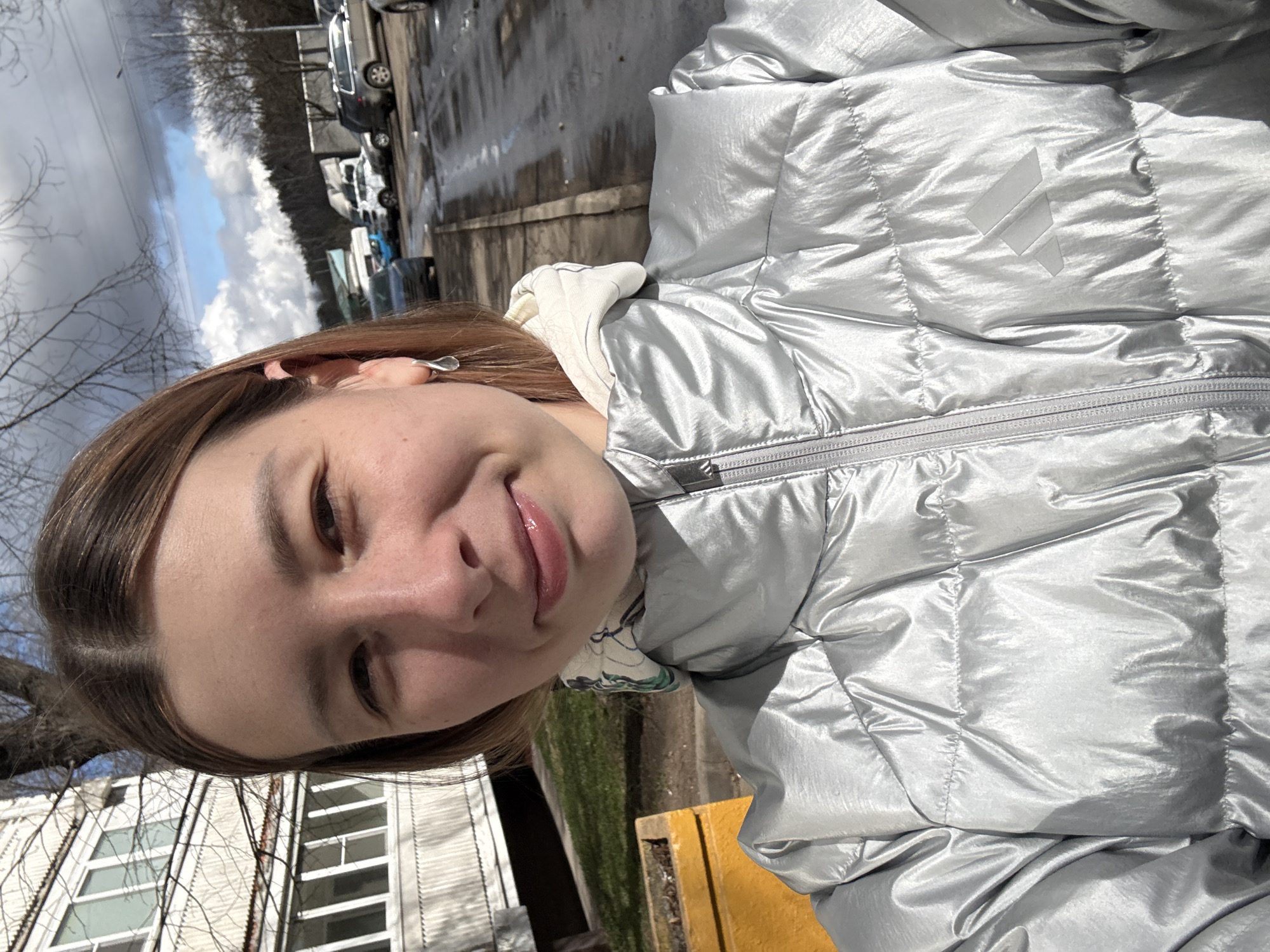 The image shows a young woman wearing a puffy silver jacket with a white hood and the Adidas logo. She is outdoors in an urban environment, as evidenced by the presence of buildings, trees, parked cars, and what appears to be a paved or asphalt surface with some wet areas. The location appears to be in Moscow, Russia. The woman is looking at the camera, smiling slightly. The weather seems to be overcast. The background includes a building with multiple windows and bare tree branches. The ground is mostly paved with some areas of grass.