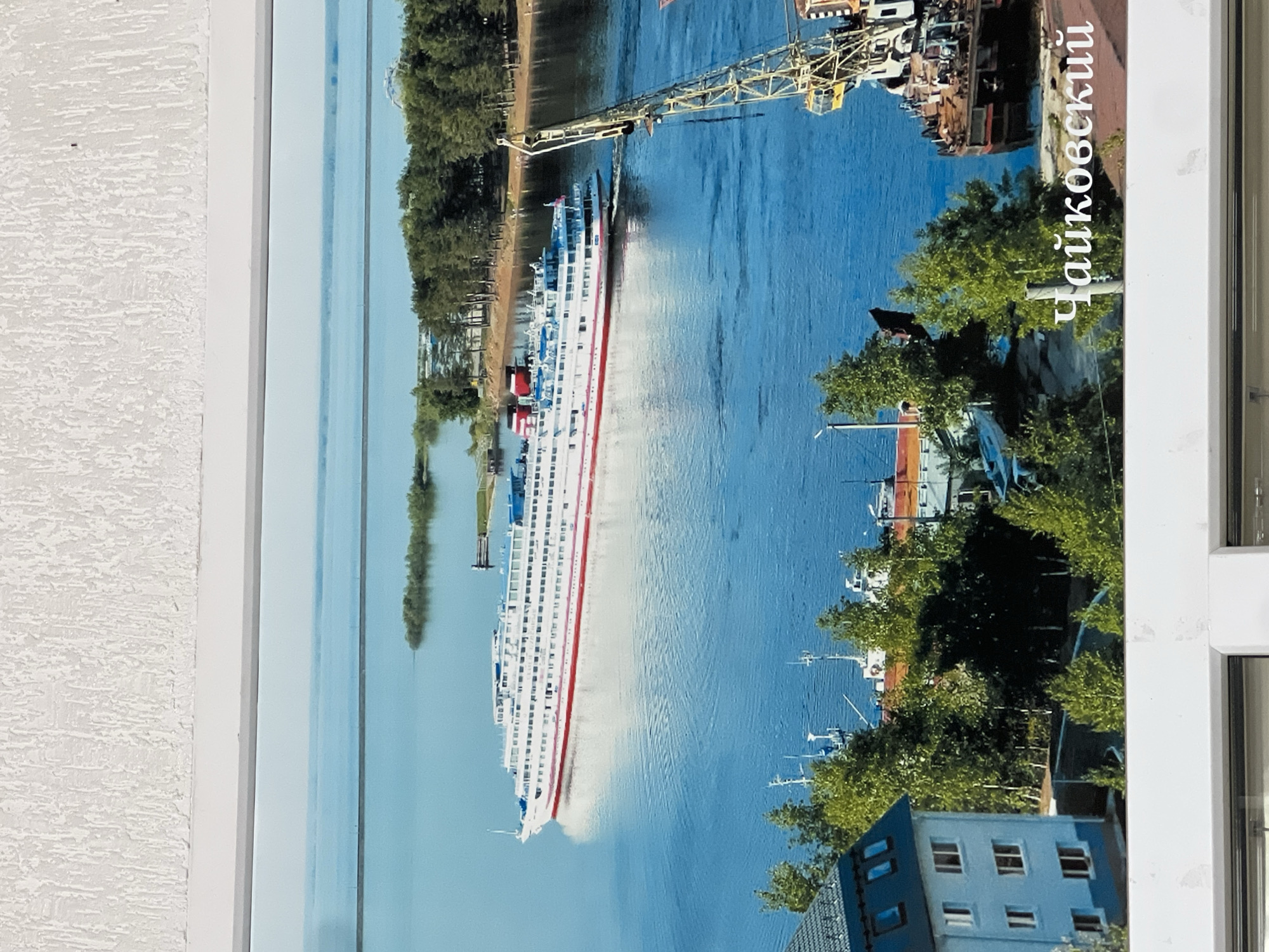 A photographic image of a cruise ship docked along a riverfront in the city of Chaikovsky, Russia is displayed on a wall. The vessel is predominantly white, with a red accent stripe and multiple decks visible. A yellow crane stands nearby, possibly involved in loading or unloading operations. The surrounding environment includes trees and buildings lining the riverbank, with a clear blue sky reflecting in the water. Text in Russian, seemingly identifying "Chaikovsky" is visible on the right side of the image. The photograph is bordered by a white frame on the left and the edge of what appears to be a window frame on the right.