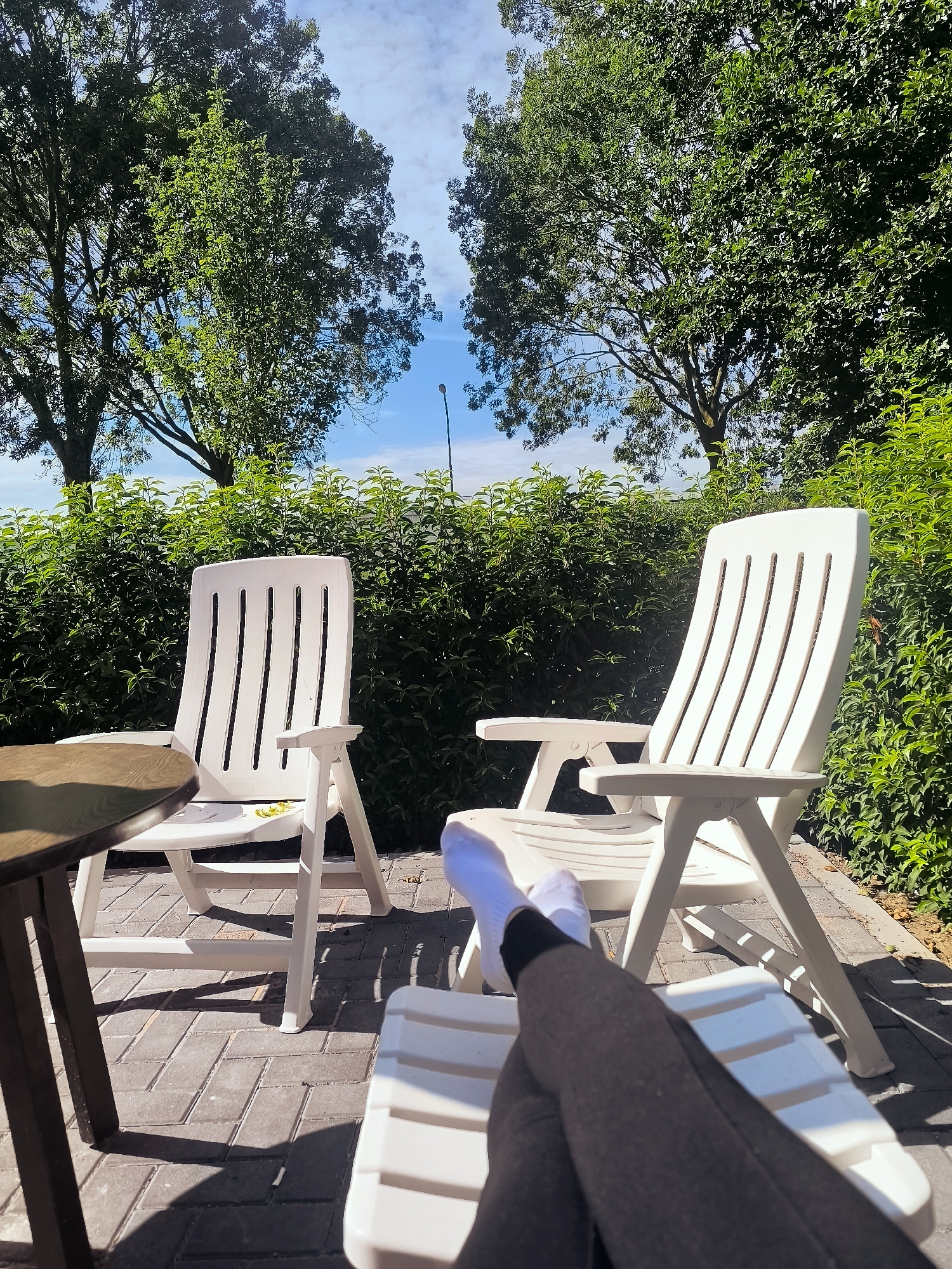 This image depicts a sun-drenched outdoor patio area, likely in Almere, Netherlands, furnished with white plastic patio chairs and a lounge chair. The ground is paved with grey interlocking bricks, and shadows cast by the furniture suggest a bright, sunny day. Lush green bushes form a backdrop, with two large trees providing shade and framing the sky. In the foreground, a person's legs are visible, clad in grey leggings and white socks, resting on the lounge chair. A small, dark wooden table is positioned to the left of the frame. The scene evokes a sense of relaxation and leisure in a pleasant outdoor setting.