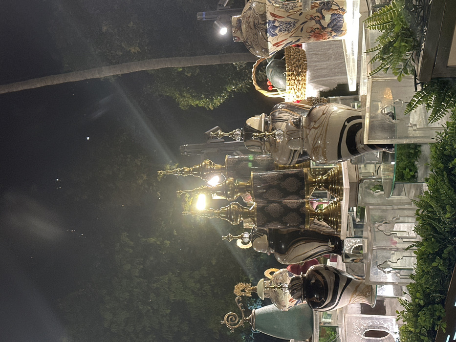 The image displays a brightly lit exhibition or retail display of decorative vases and home decor items against a dark, nighttime outdoor background. The photograph is oriented 90 degrees counter-clockwise from a standard upright view.
In the foreground, a multi-tiered display showcases numerous ornate vases and jars. The items are arranged on clear, possibly acrylic or glass, shelving units, some of which feature decorative cut-out patterns. To the far right, lush green foliage, resembling ferns, adds a touch of natural accent to the display.
The decorative pieces themselves vary greatly in style and material. Towards the upper right, a large, intricately patterned ceramic vase features a blue and orange floral design. Below this, and continuing across the display, are rows of distinct vases. Many are characterized by glossy finishes, with patterns such as black and white stripes, mottled marble-like effects, and geometric designs on darker, cylindrical bodies. The majority of these vases incorporate metallic, gold-colored bases, necks, and decorative finials. Several of the finials are adorned with clear, crystal-like elements, adding to their luxurious appearance. One unique piece towards the bottom left of the display is a teal or greenish vase with an elaborate golden, swirling handle or embellishment. A woven basket is also subtly visible behind some of the vases in the upper right section.
The display is illuminated by multiple warm, yellow artificial lights, some of which are visible as bright glows or cause significant light streaks and reflections, particularly on the shiny surfaces of the vases and metallic elements. These lights create a vibrant and inviting atmosphere for the exhibited items.
The background is predominantly dark, indicating that the scene takes place at night. Dark silhouettes of trees are visible, with one prominent, slender tree trunk (likely a palm) extending diagonally across the upper left portion of the image. Scattered within the dark background are several small, faint white lights, which could be distant light sources or atmospheric phenomena.
The overall impression is one of an elegant nighttime display, possibly at a market, outdoor shop, or special event, showcasing ornate decorative objects. No visible text is present in the image.