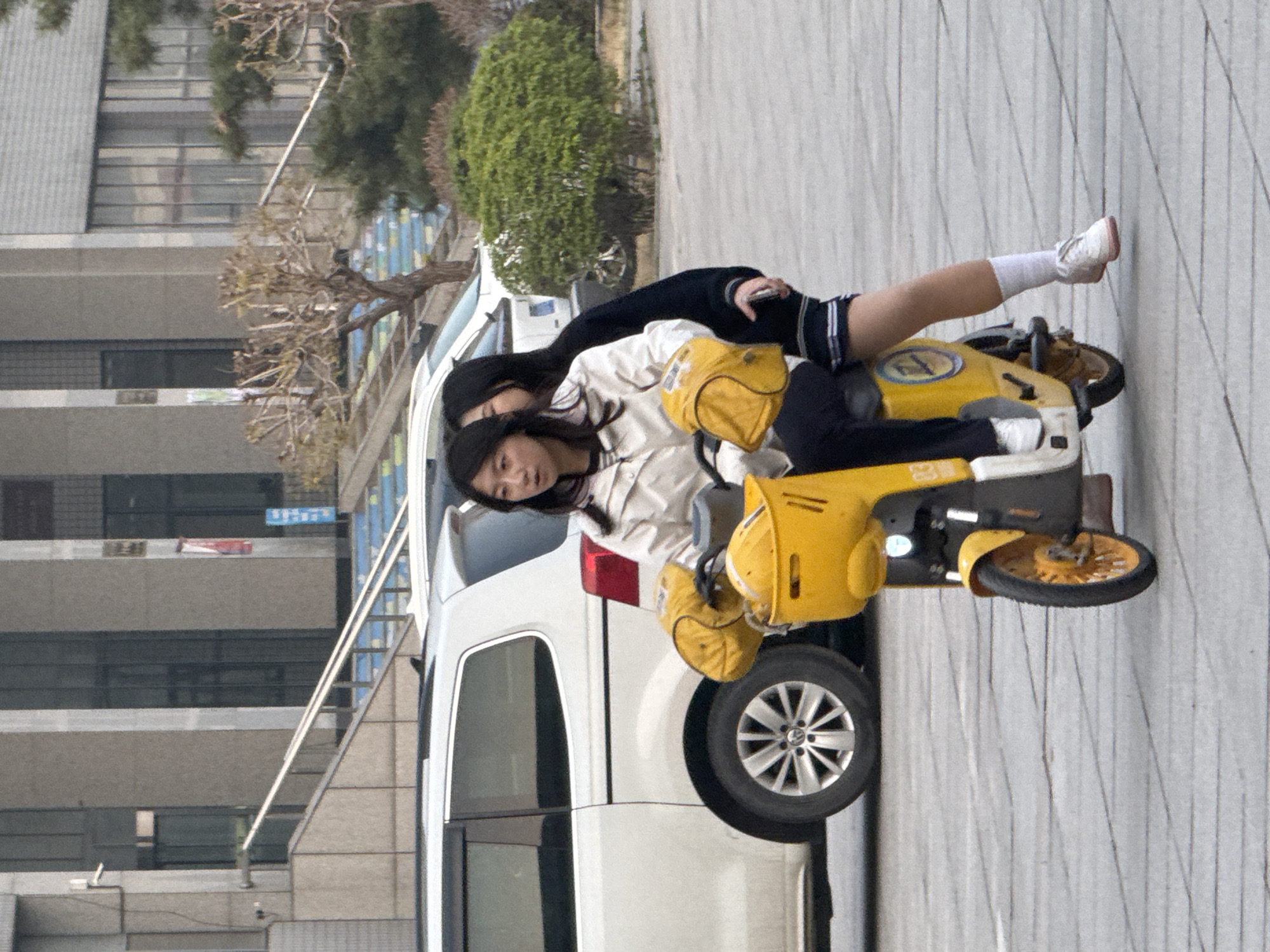 Two females are on a yellow motorized scooter next to a white van on a paved surface. One female is operating the scooter, while the other is seated behind. The location appears to be an urban setting in Zhongtun, China, indicated by the architecture and environment. A multi-story building is in the background, with other greenery and a street.