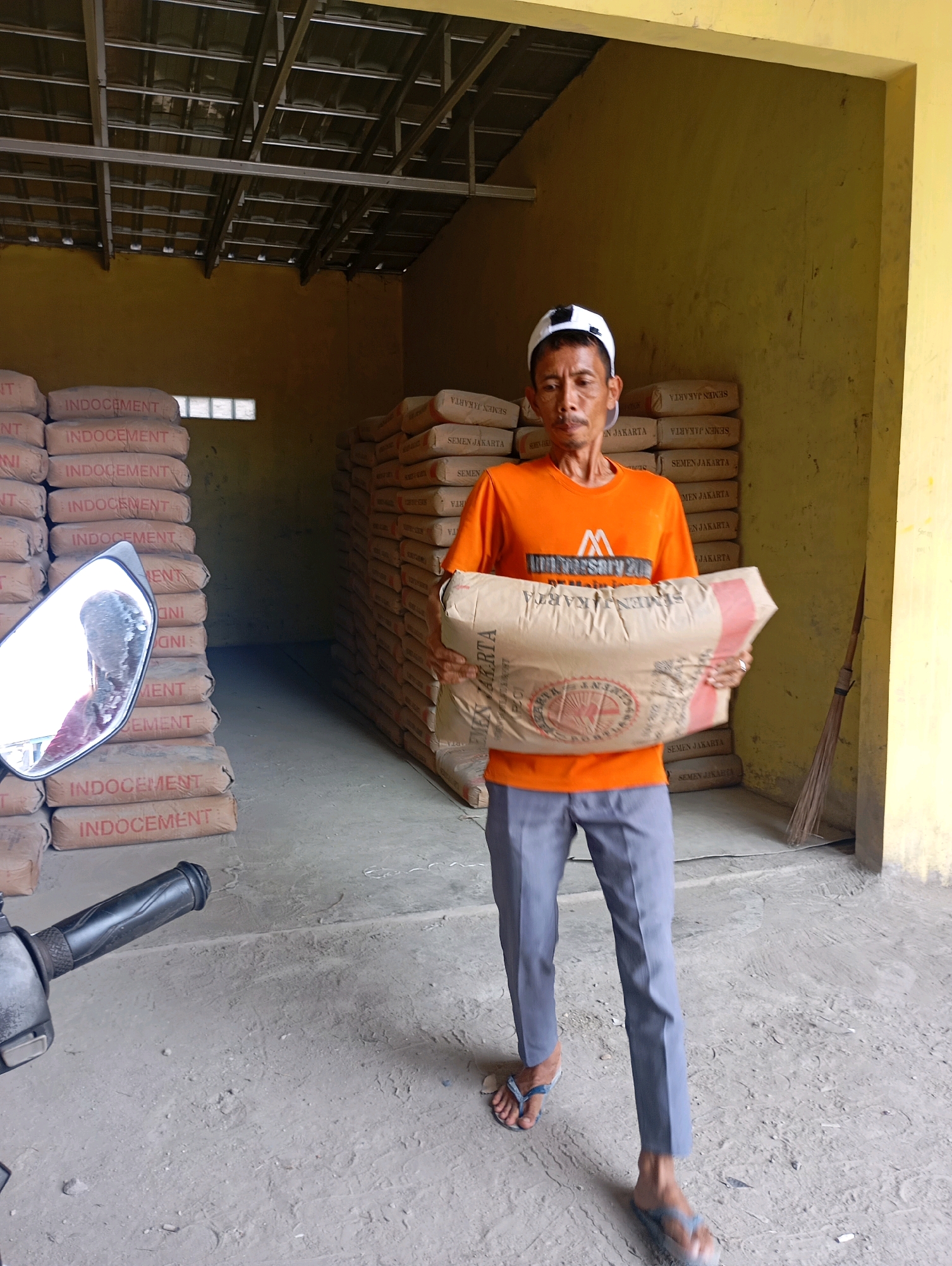 A man in an orange t-shirt and gray pants is standing in a warehouse with stacks of cement bags. He is holding a large bag of cement. The warehouse appears to be in Ciakar, Indonesia, as suggested by the text on the cement bags. The floor is concrete, and the walls are painted yellow. A broom leans against the wall on the right side of the image. A motorcycle is partially visible on the left side of the image. The roof of the warehouse is made of metal.