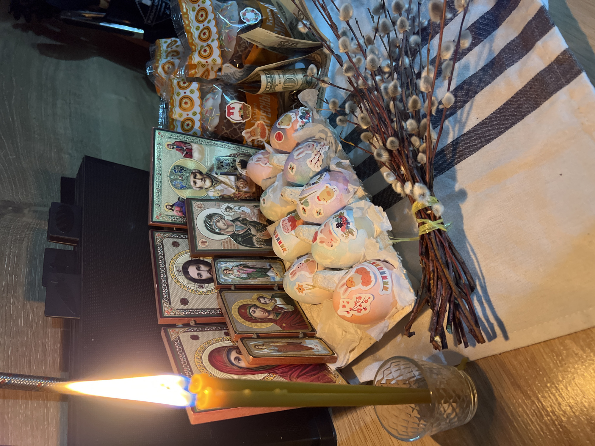 A still life composition on a table, likely indoors, in Kazan, Russia, exhibits elements of an Orthodox Christian Easter celebration. The scene includes a lit candle in a glass holder, several religious icons, decorated Easter eggs, pussy willow branches tied with yellow tape, packaged Easter bread (kulich), and a folded U.S. dollar bill. The table is partially covered with a white cloth with dark diagonal stripes. The scene appears to be on a table or surface.