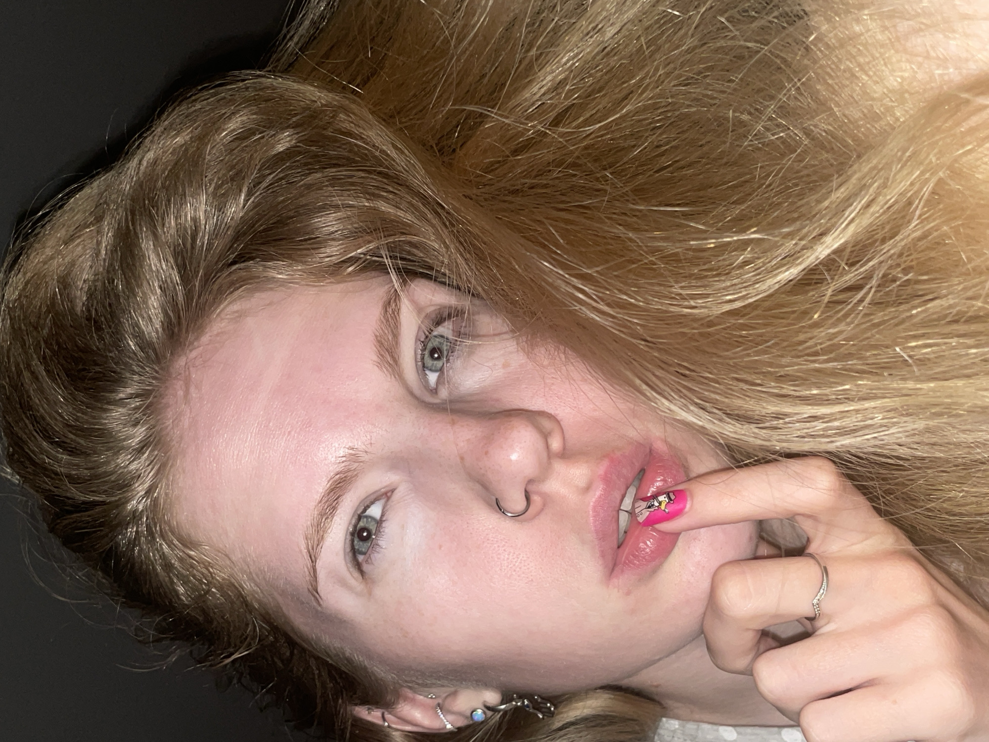 A young woman with long, blonde hair is lying down, facing the camera. She has light skin, blue eyes, and a silver nose ring. Her finger is near her mouth, revealing a pink nail with a cartoon design. She is wearing a silver ring on her finger and earrings in her ear. The background is dark and blurry. The location is not discernible from the image.