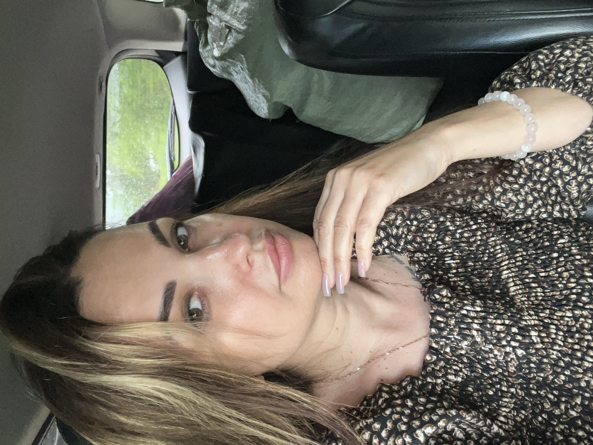 A woman is pictured from the chest up, looking sideways with her chin resting on her hand. She has long, wavy brown hair with blonde highlights. She is wearing a brown and white patterned top and a delicate gold necklace. A bracelet with light pink beads is visible on her right wrist. The background suggests she is in the back seat of a vehicle, with a view of green foliage through a rear window, indicating an outdoor setting. There are large green bags in the backseat behind her. The lighting suggests it is daytime, possibly overcast.