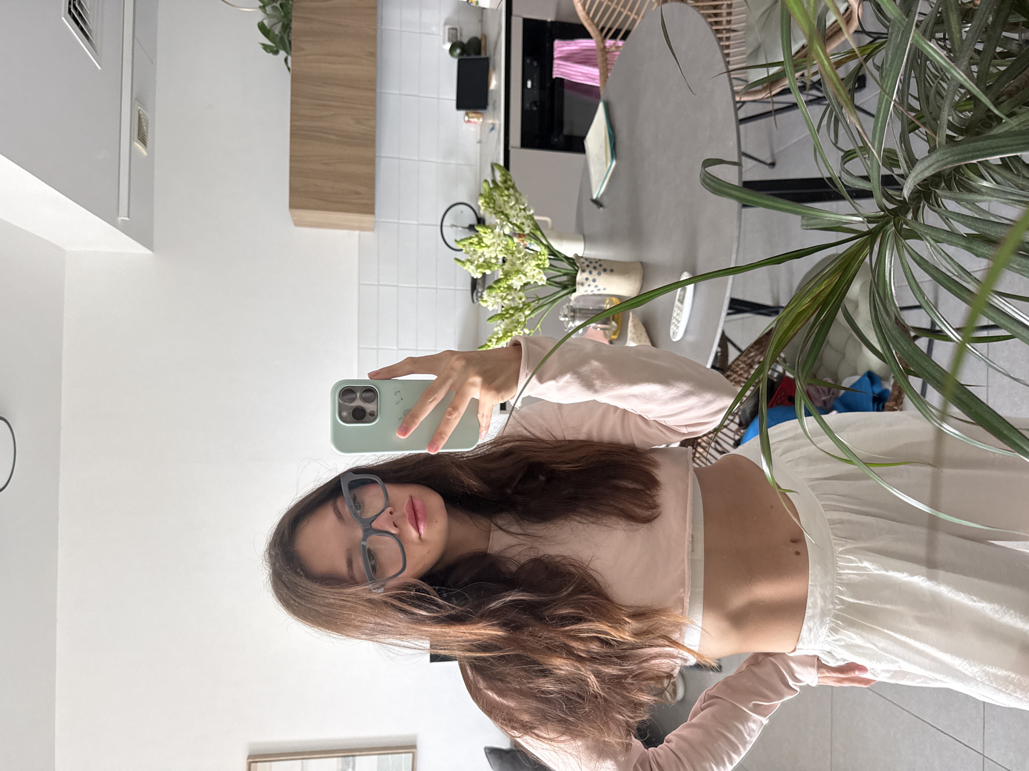 A woman stands in a brightly lit kitchen, taking a selfie in a mirror. She has long, wavy brown hair and is wearing light blue-rimmed glasses, a pink crop top, and white wide-leg pants. Her midriff is exposed. In the background, a gray round dining table is visible, adorned with a vase of white flowers. To the right of the table, a wicker chair and a portion of a dining area are visible. Further back, kitchen cabinets, a stovetop, and a clock are present. A green leafy plant stands in the foreground, partially obscuring the right side of the frame.