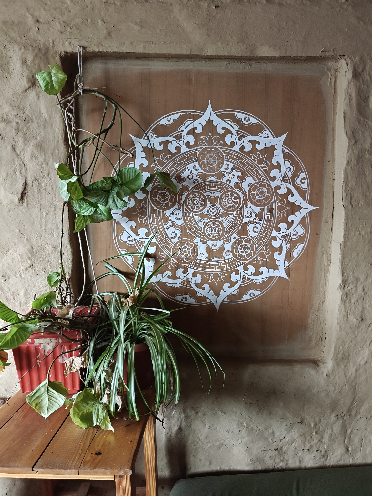 In Prini, India, a close-up shot captures a detail of a wall adorned with a white mandala pattern on a brown wooden panel. To the left, a red planter holds two types of plants: a vine with heart-shaped leaves cascading down and a spider plant with long, striped leaves. The wall itself is textured, appearing to be made of rough plaster or adobe. A wooden table sits in the foreground, supporting the planter. The overall impression is one of rustic, natural decor.