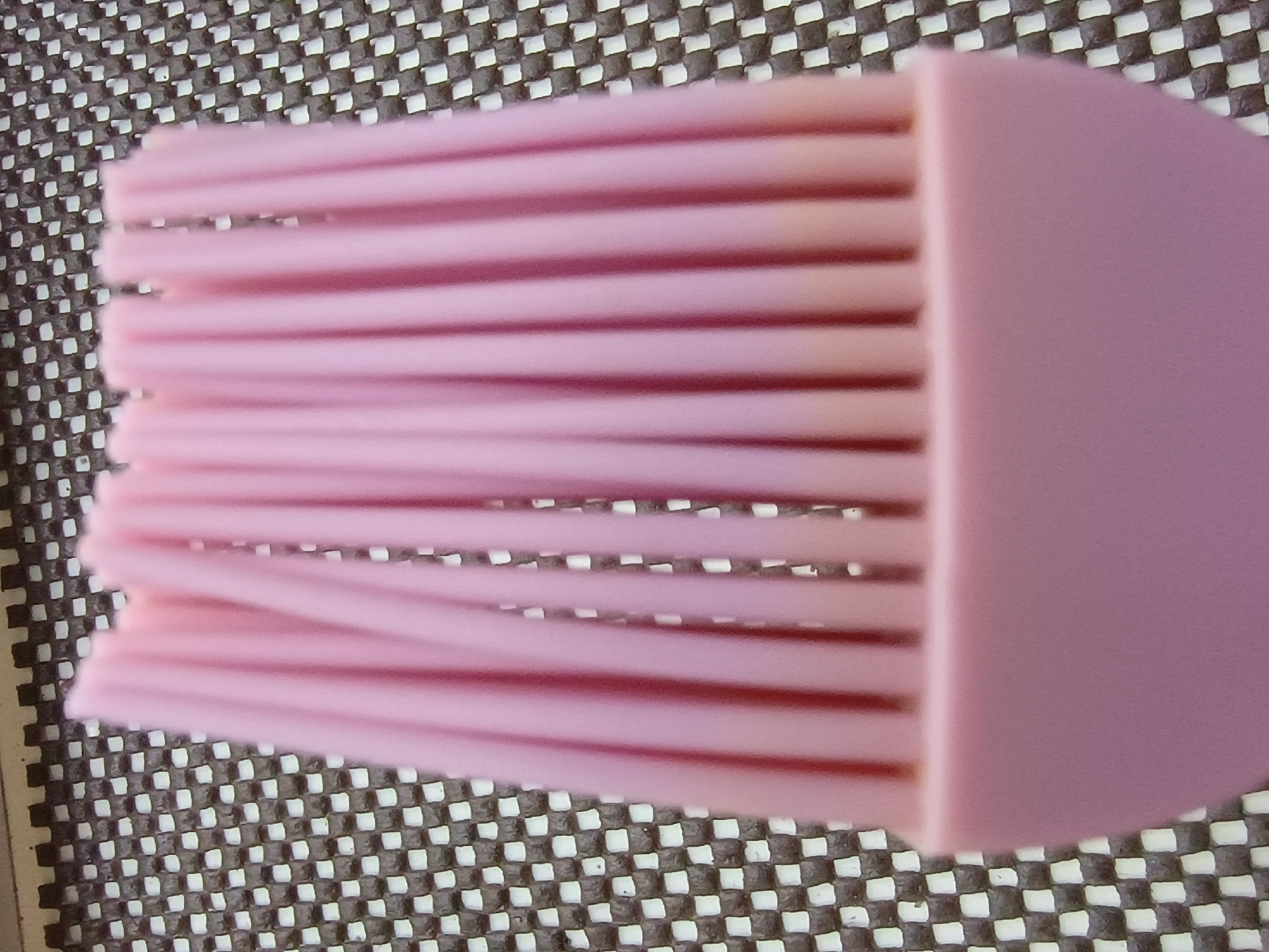 A close-up, eye-level shot shows a pink silicone pastry brush resting on a dark, textured surface with a checkered pattern of white and brown squares. The brush is oriented diagonally from the bottom left to the top right of the frame, with its bristles dominating the majority of the image. The bristles are thin, flexible, and uniformly spaced, creating a ribbed effect. They are a vibrant pink color, with some appearing slightly darker than others due to lighting. The handle of the brush, also pink, is visible on the right side of the frame, appearing solid and smooth, with a rounded edge. The background surface is out of focus, but its distinct checkered pattern suggests a placemat or a textured tabletop. The lighting appears to be neutral, casting soft shadows that highlight the texture of the bristles. There is no text visible in the image. The scene is static, focusing solely on the object. The setting is likely domestic or a kitchen environment given the nature of the object. The image does not convey a specific time of day, weather, or emotional state.