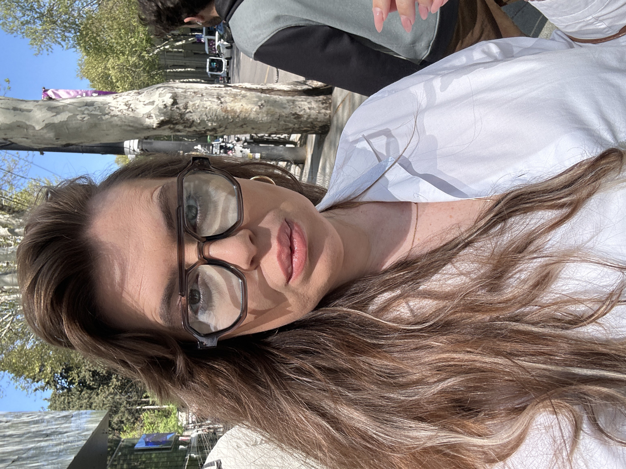 A woman with long brown hair and glasses poses for a selfie. She wears a white shirt and gold earrings. Behind her is a person partially visible in a grey and black jacket. The background shows a tree-lined street with buildings and vehicles. This image was taken in Tbilisi, Georgia.
