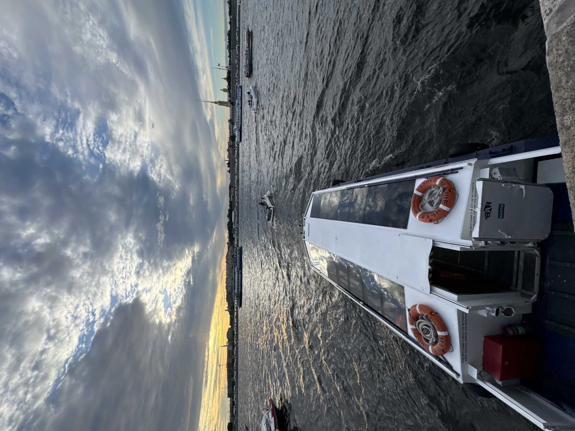The image captures a view from a boat on a river in Saint Petersburg, Russia, likely during the late afternoon or early evening given the warm light. In the foreground, the stern of a white and blue passenger boat is visible, with two orange life preservers prominently displayed. Red and white lettering on the boat reads, "ПОД МОСТАМИ С МЕСТ НЕ ВСТАВАТЬ KEEP SEATED UNDER THE BRIDGES." Further in the distance, across the choppy, dark water, a cityscape unfolds. Tall spires, characteristic of Saint Petersburg architecture, rise above the buildings lining the riverbank. Several other boats, including a larger tour boat, are navigating the waterway. The sky above is dramatic, with a mix of heavy gray clouds and patches of blue, hinting at the possibility of an approaching storm or the aftermath of one, with the sun breaking through in a golden band on the horizon. The overall atmosphere is one of urban river life under a dynamic sky.