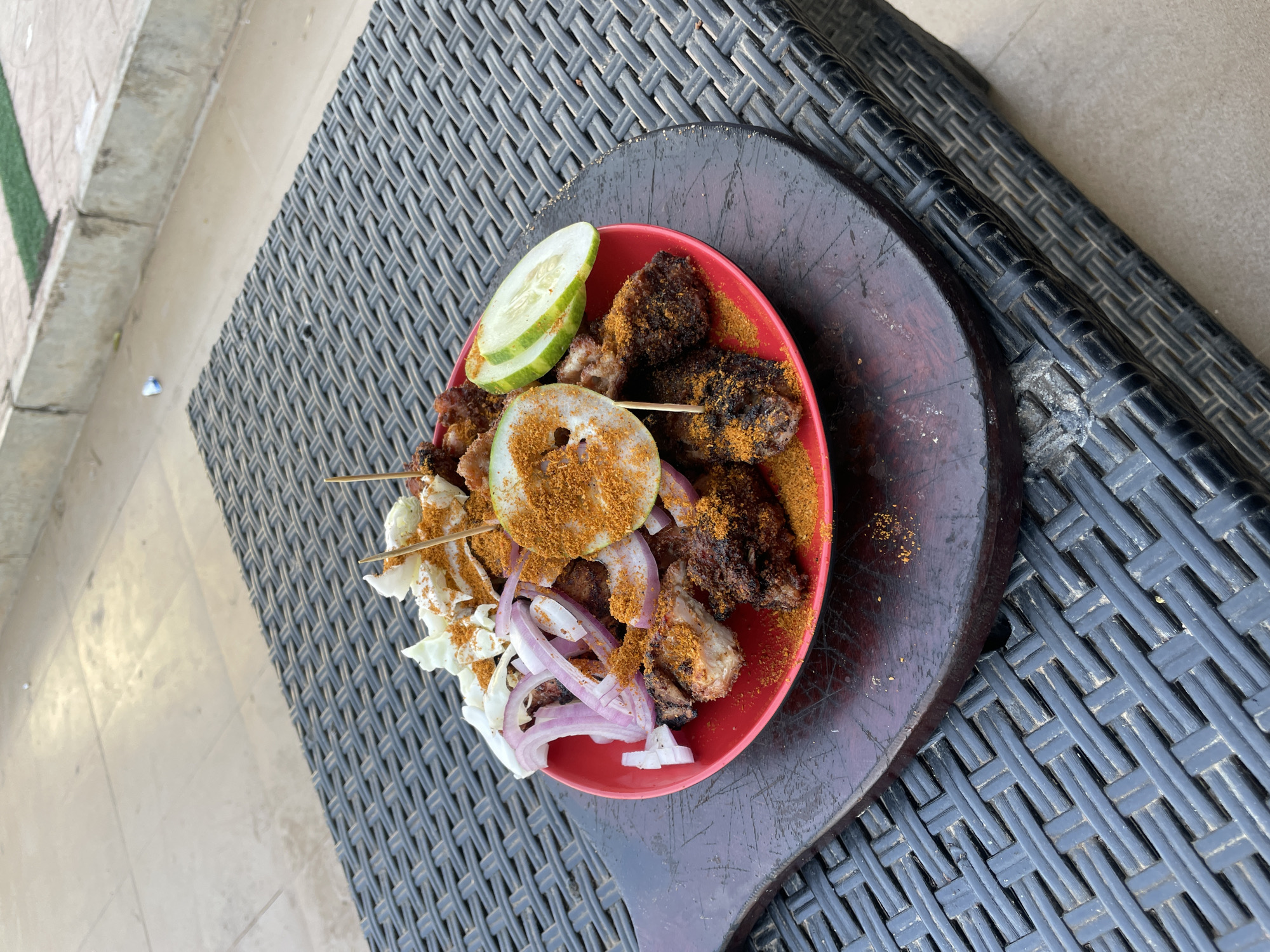 A plate of suya, a spicy grilled meat skewer popular in Nigeria, sits on a dark wooden platter. The platter is placed on a black wicker chair or table. The suya is presented on a red plate and includes skewers of grilled meat, sliced red onions, shredded cabbage, and cucumber slices. A sprinkling of a reddish-brown spice is evident on the dish. The setting appears to be outdoors, possibly near a pool or patio area given the presence of nearby flooring. The location is reported as Barakin Lerawi, Nigeria.