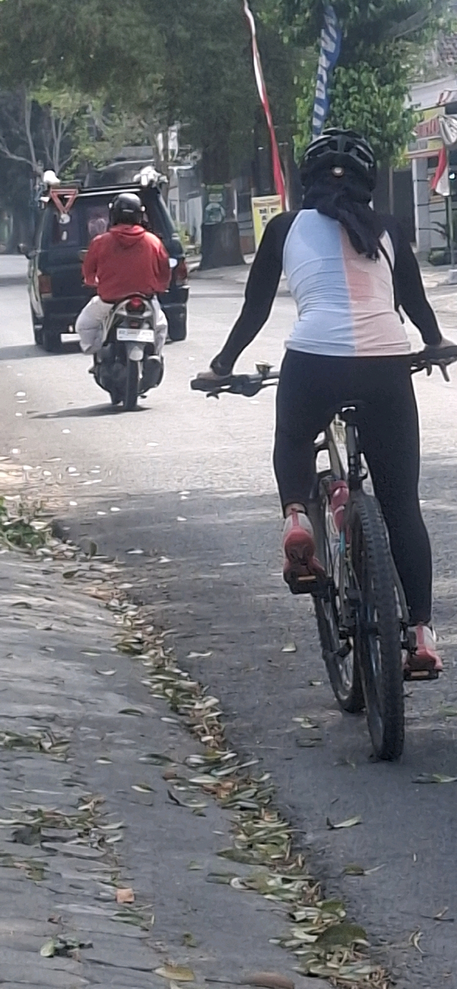 In Jiwut, Indonesia, a medium-shot vertical image captures two individuals on a road. On the left, a person wearing a red hoodie and white pants rides a scooter, facing away from the camera. A black vehicle is positioned behind them, with part of its roof visible. On the right, another individual, seen from behind, cycles on a bicycle, also facing away. This cyclist wears a helmet, a long-sleeved jersey with pink and light blue panels on the upper torso, black leggings, and red athletic shoes. The road surface is asphalt and is strewn with fallen leaves. In the background, trees line the road, and buildings are visible, suggesting an urban or suburban setting. Flags and poles are also present in the background.