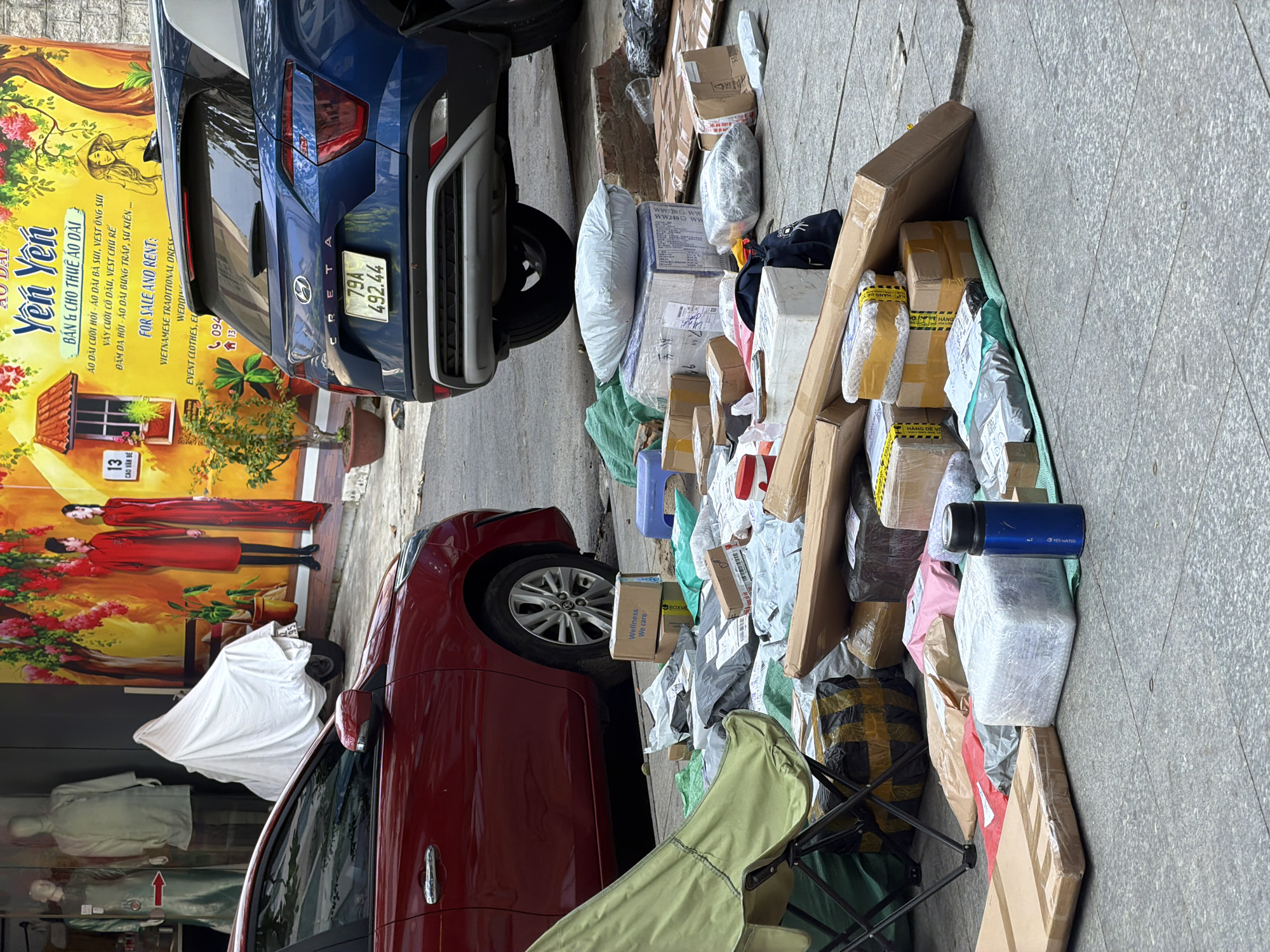 The image depicts a street scene in Bac Nha Trang Ward, Vietnam, with a large accumulation of packages on the sidewalk. A red car is parked in the foreground, partially obscuring a collection of parcels. Behind the red car, a blue SUV is also visible. The packages vary in size and are wrapped in brown paper, plastic, and some are in cardboard boxes. Some packages are secured with tape. A green foldable chair is positioned to the left of the frame, near the red car. A blue water bottle is placed on top of some of the packages. In the background, there is a shop with mannequins displaying clothing, and a large yellow banner with Vietnamese text and imagery. The ground is paved with stone slabs.