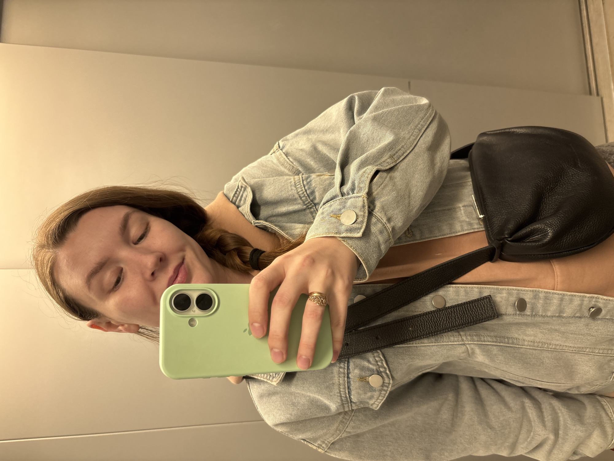 A young woman takes a selfie indoors with a light green phone. She wears a light blue denim jacket over a peach-colored top, with a black leather bag slung across her body. Her brown hair is styled with a braid. A ring adorns the ring finger of her left hand. The background is a plain, light-colored wall. No identifying features point to Izhevsk, Russia, or any specific city.