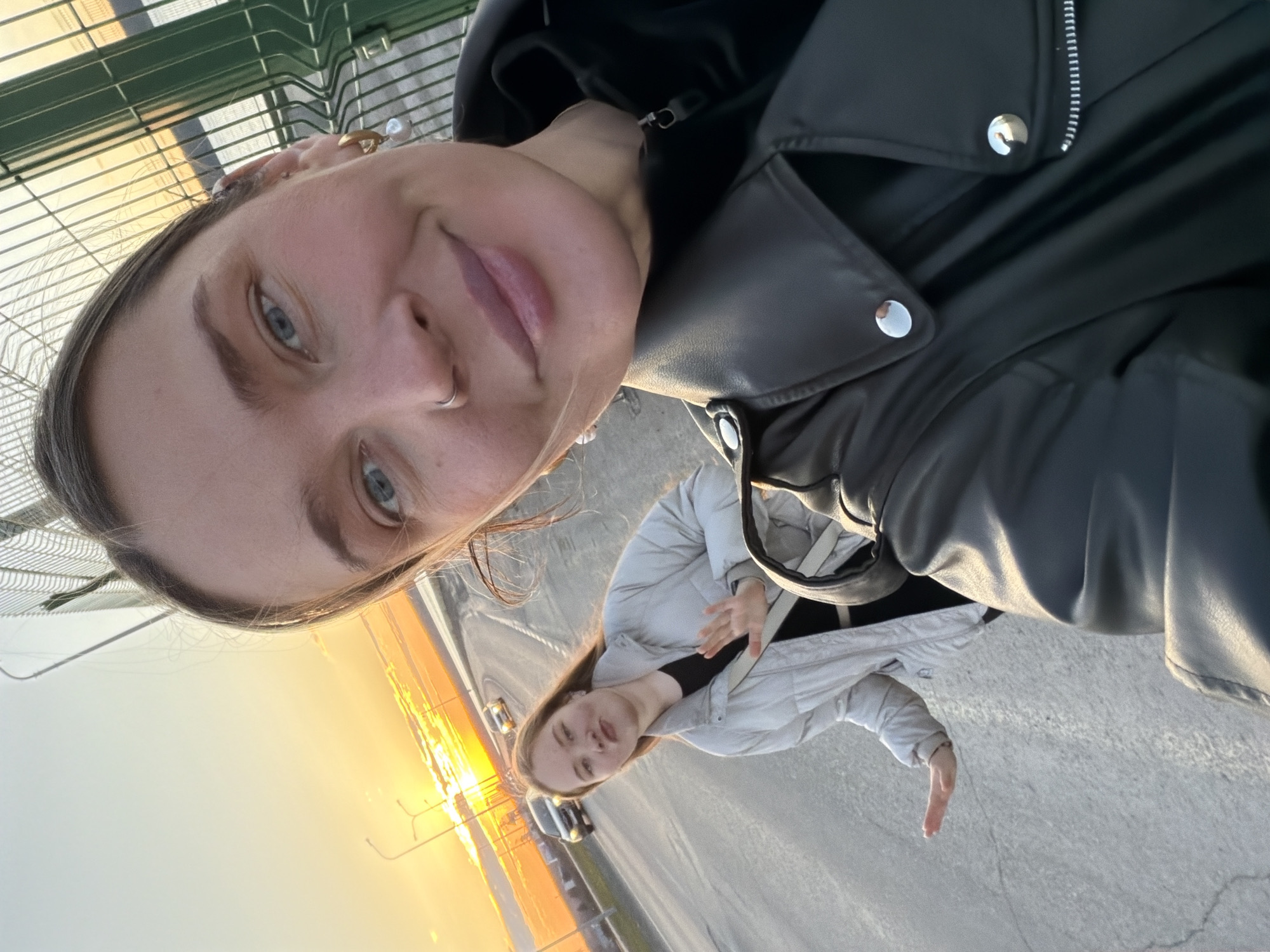The image shows a selfie taken by a woman, likely in her 20s or 30s, wearing a black leather jacket with silver buttons. She has fair skin, blue eyes, and a nose ring. Behind her, another woman stands near what appears to be a highway or major road during sunset. This second woman wears a light-colored puffer jacket.
The location is likely Chaikovsky, Russia, inferred from the metadata. The background shows infrastructure consistent with a roadway, including street lights and vehicles. A fence is visible in the upper left corner of the image. The time of day is near sunset, indicated by the bright orange and yellow light in the background.