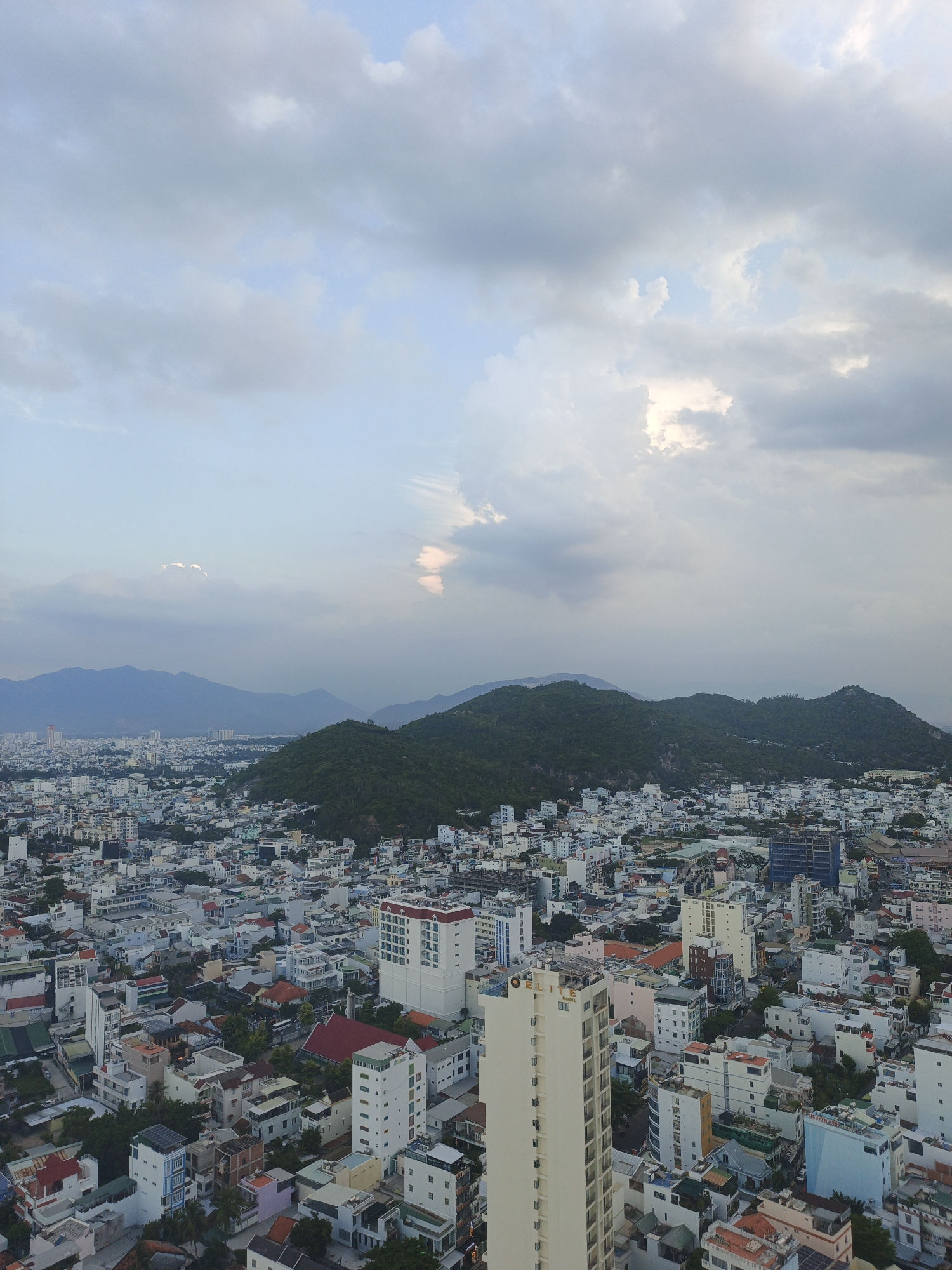 This is a high-angle aerial photograph of the city of Nha Trang, Vietnam, specifically the Bac Nha Trang Ward, captured during daylight hours under a cloudy sky. The foreground is dominated by a dense urban landscape featuring numerous buildings of varying heights and architectural styles, predominantly in white and light colors, with some accented by red or blue roofs. Narrow streets are visible winding between the structures. In the mid-ground, a prominent, light-colored tall building stands out, possibly a hotel, with the "ELITE HOTEL" signage visible. Further in the background, a range of verdant, tree-covered hills forms a backdrop to the city. The sky is filled with dramatic, grey and white clouds, with patches of blue sky visible in between, and a bright area of light suggests the sun may be breaking through in the distance. No people are discernible in the image.
