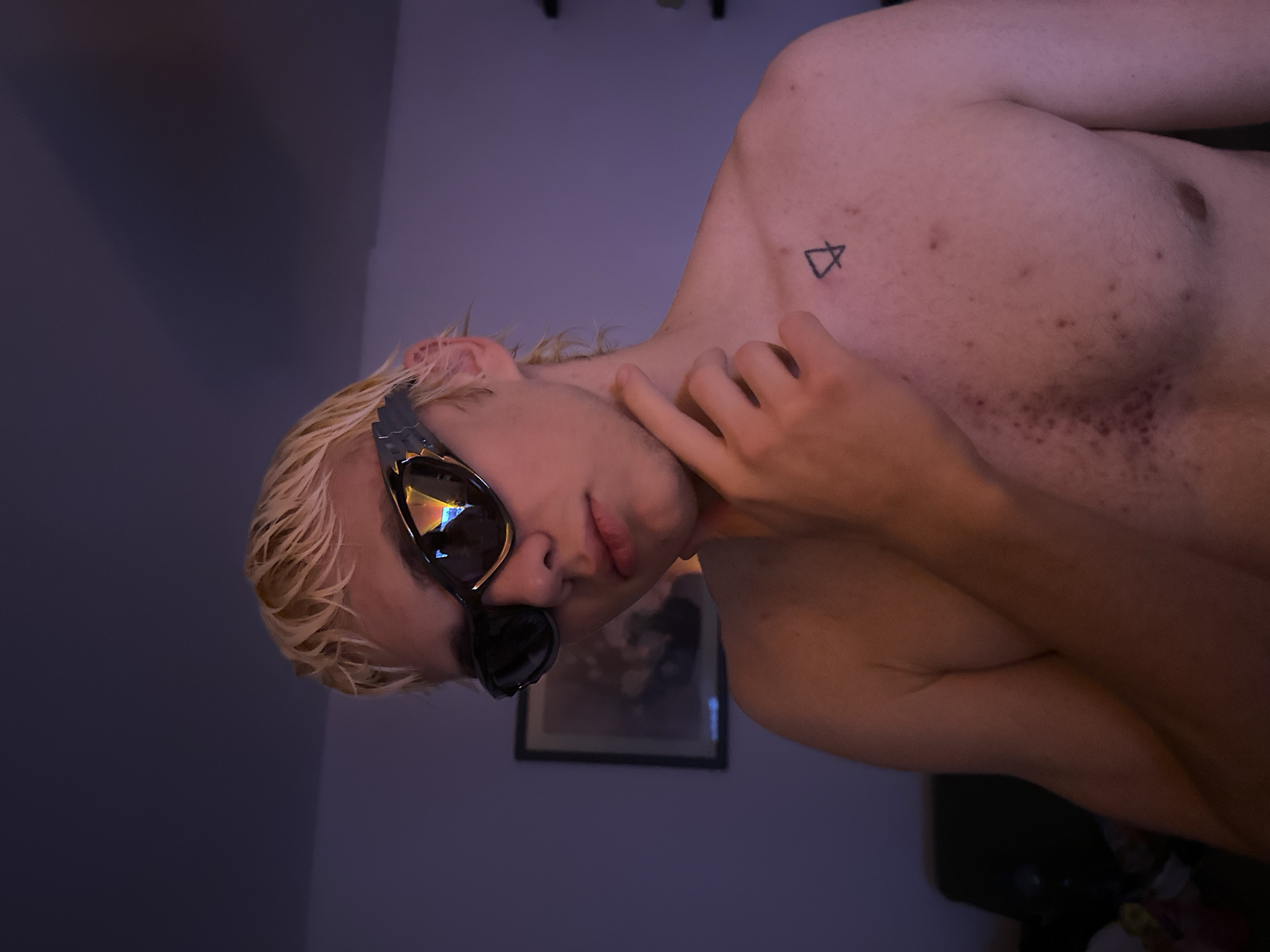 A shirtless, fair-skinned individual with blonde hair and sunglasses poses for the camera. They have a small triangular tattoo on their left shoulder and are touching their neck with their right hand. A framed picture hangs on the wall behind them. The location is unknown, and there is no information indicating the city of Novi Sad, Serbia.