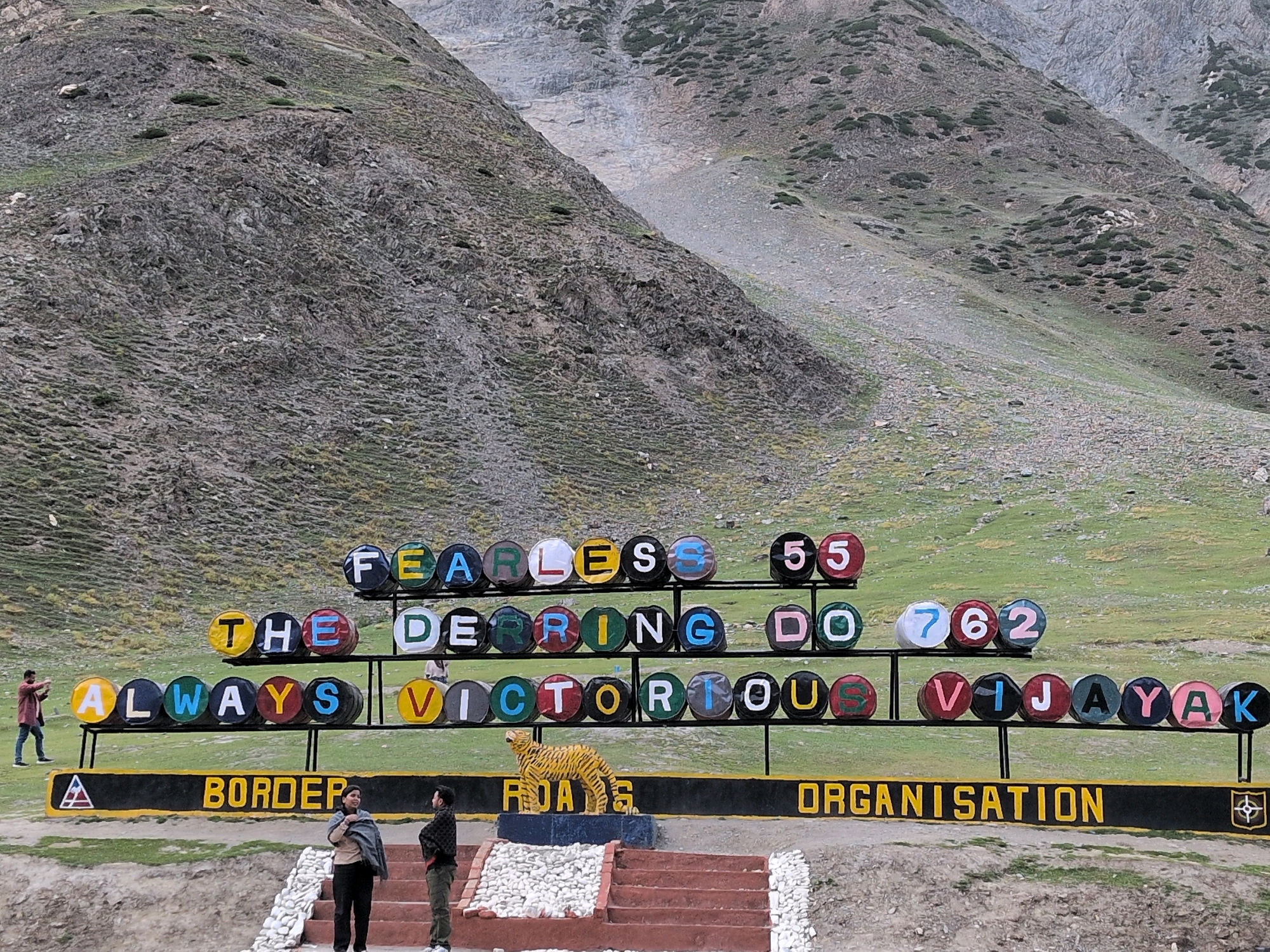 An outdoor scenic shot captures a mountainous landscape with a sign that reads "FEARLESS 55 DERRING DO 762 ALWHYS VICTORIOUS VIJAYAK BORDER ROADS ORGANISATION". The sign is made of colorful barrels and is mounted on black poles. The background features rolling green hills and rocky mountains, indicating a high-altitude location. In the foreground, two individuals stand on a set of stairs, observing the sign. The person on the left is wearing a black shirt and green pants, while the person on the right is wearing a beige shawl and black pants. Another individual is visible in the distance on the left, taking a photograph. A tiger statue is positioned in front of the "BORDER ROADS ORGANISATION" part of the sign.