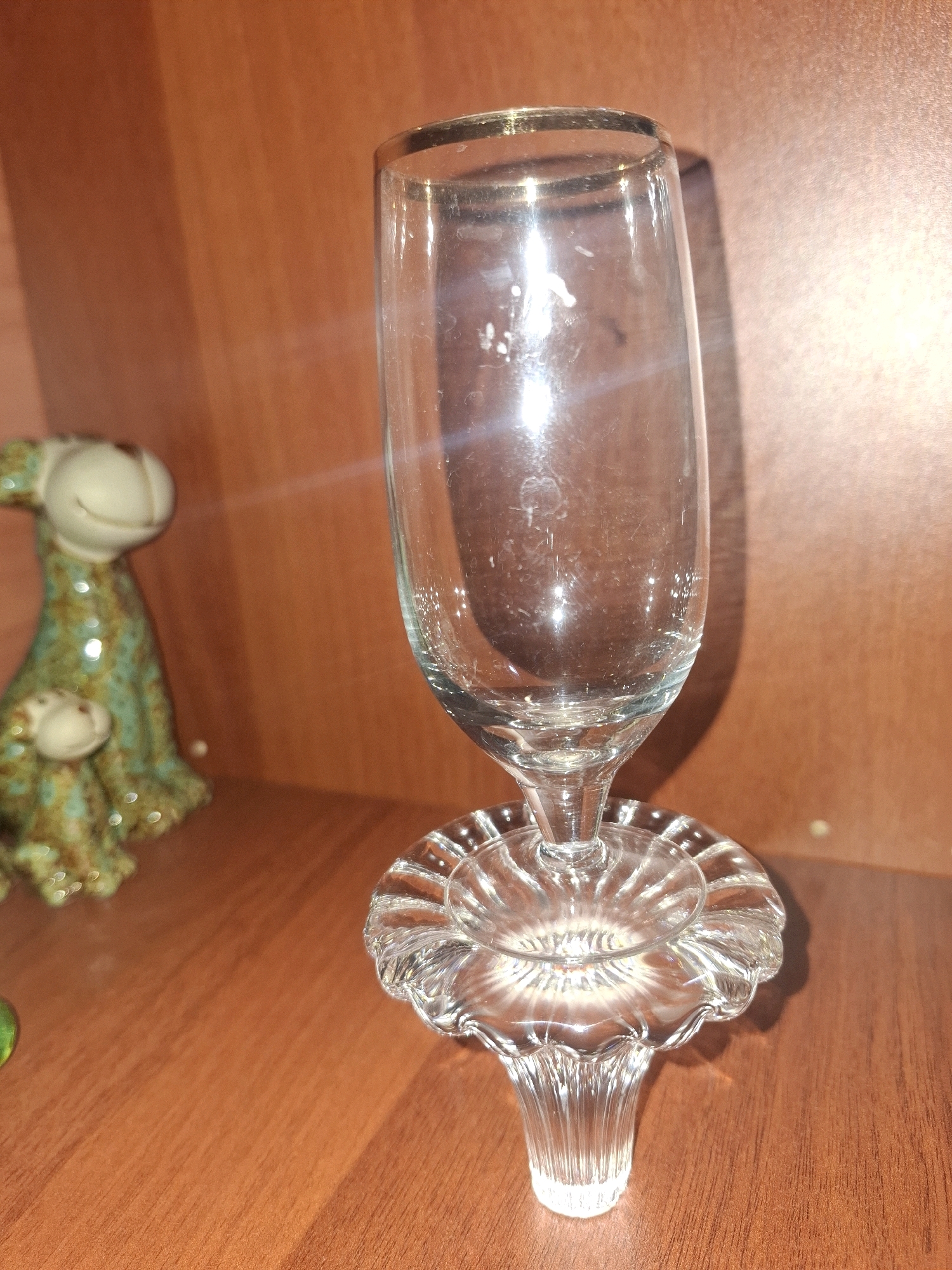 A clear glass champagne flute with a decorative glass base stands on a wooden surface. To the left, a small, green, speckled ceramic figurine with a smiling face is visible. The background is a textured, orange-brown surface, possibly a shelf or wall. The lighting creates reflections on the glass and highlights the textures of the objects. The scene appears to be indoors, possibly on a shelf in a home or shop. There is no discernible indication of the location being in Almaty, Kazakhstan.