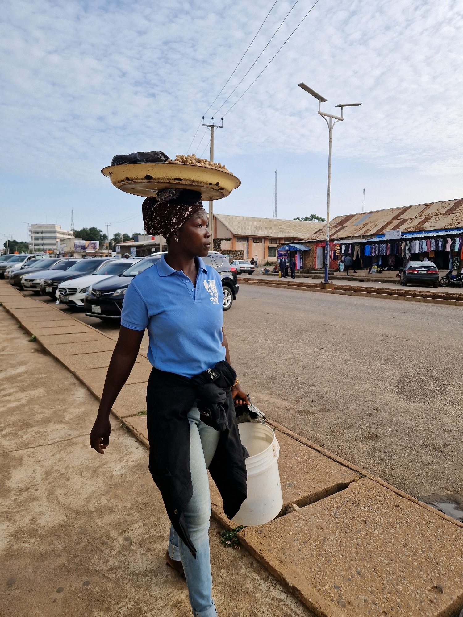 A woman walks on a sidewalk carrying a tray of food on her head. She is wearing a blue polo shirt, jeans, and a headscarf. She is also carrying a white bucket in her right hand. The sidewalk is made of concrete and is next to a road with several cars parked along the side. In the background, there are buildings and shops. The sky is cloudy.