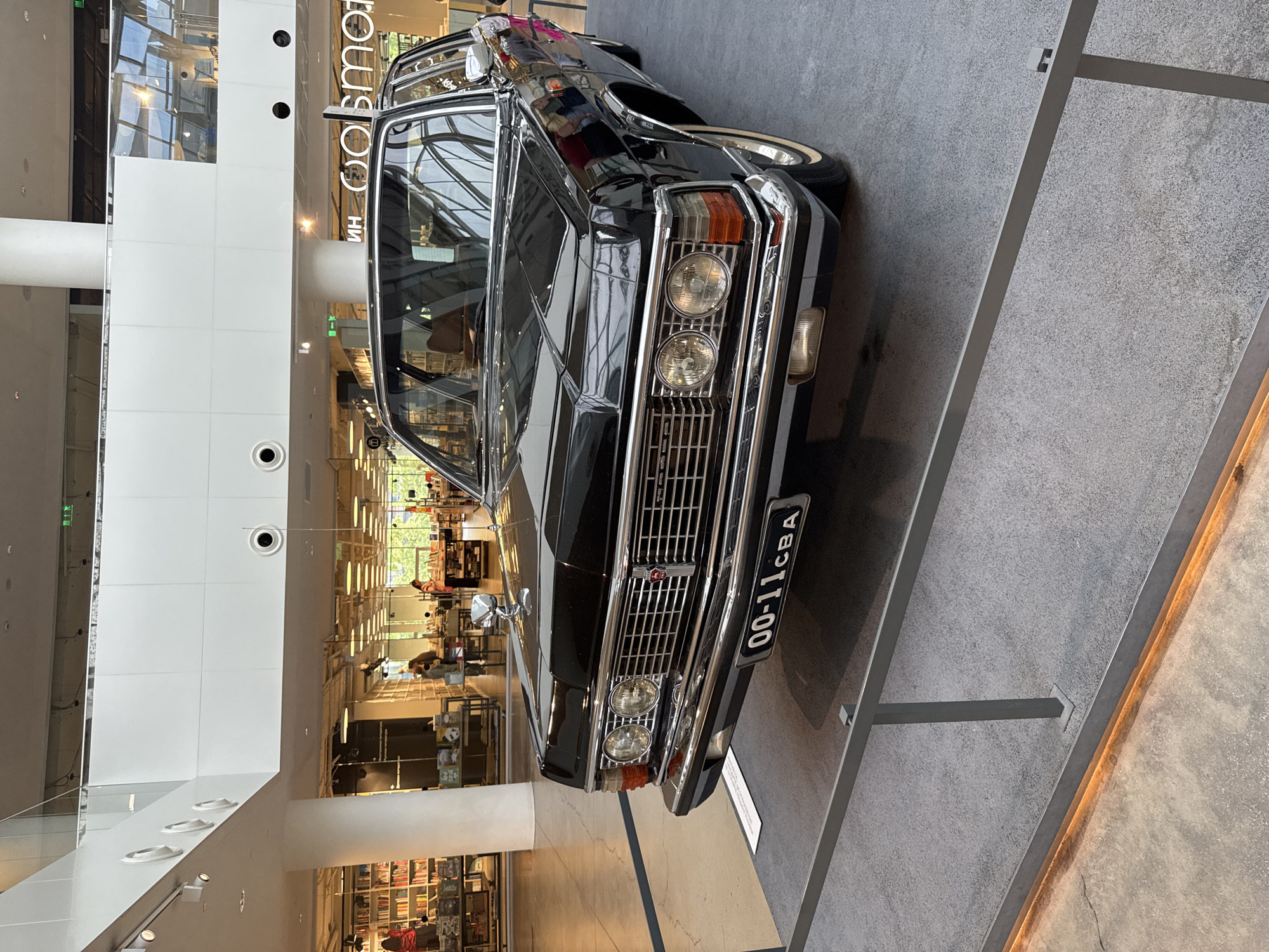 This media file showcases a black vintage sedan, likely a ZIL, displayed in an indoor exhibition. The car is positioned at an angle, with its front grille and headlights prominently visible. The license plate reads "00-11CBA". The car is on a raised platform with a grey, textured surface.
The background reveals a modern, multi-level interior with large windows allowing natural light to filter in. The architecture features white walls, columns, and what appear to be escalators or walkways. In the distance, through the windows and open spaces, there are glimpses of people browsing in what resembles a store or a museum gift shop, with shelves stocked with books and other items. The overall ambiance suggests a public exhibition or a shopping mall.
Visible text includes "ИН СОБМОТ" on a sign in the background. The time of day appears to be daytime, with ample lighting, and the weather is not discernible. The scene is static, indicating the car is on display rather than in motion. The setting is likely an exhibition hall or a retail space in Yekaterinburg, Russia, given the location context.