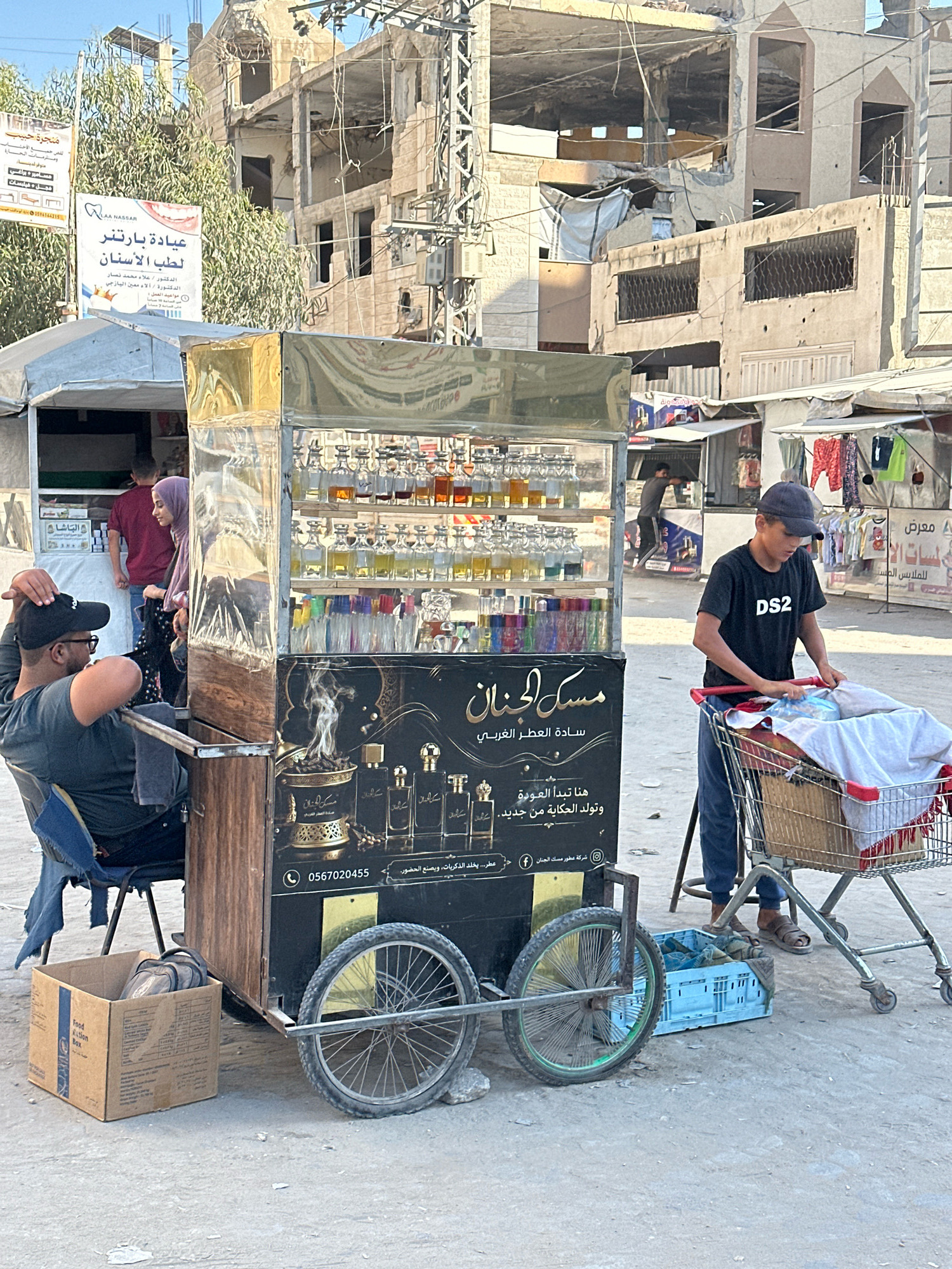 In the city of Gaza, Palestinian Territories, a street scene unfolds with a focus on a perfume cart and its surroundings. The cart, a prominent structure, features shelves stocked with numerous glass bottles of various sizes and colors, presumably containing perfumes or oils. A large sign on the cart displays Arabic text and images, including a depiction of incense burning. Two wheels, one larger than the other, are attached to the cart, suggesting it's mobile.
In front of the cart, a man with a black cap and glasses is seated in a chair, looking towards the cart. To his left, a cardboard box labeled "Food Box" rests on the ground. Nearby, a young boy wearing a dark t-shirt with "DS2" printed on it and a dark cap is pushing a shopping cart filled with clothing.
The background of the image reveals the context of the location. Several damaged buildings, appearing to be under construction or reconstruction, dominate the skyline. These structures show signs of damage, with exposed concrete and incomplete walls. Signs in Arabic are visible on some of the buildings, including a poster for a dental clinic. Clothes are also displayed on racks in the background, suggesting a market area. The ground is paved with a rough, light-colored surface, characteristic of a street or open area. The overall atmosphere suggests a bustling, if somewhat precarious, urban environment.