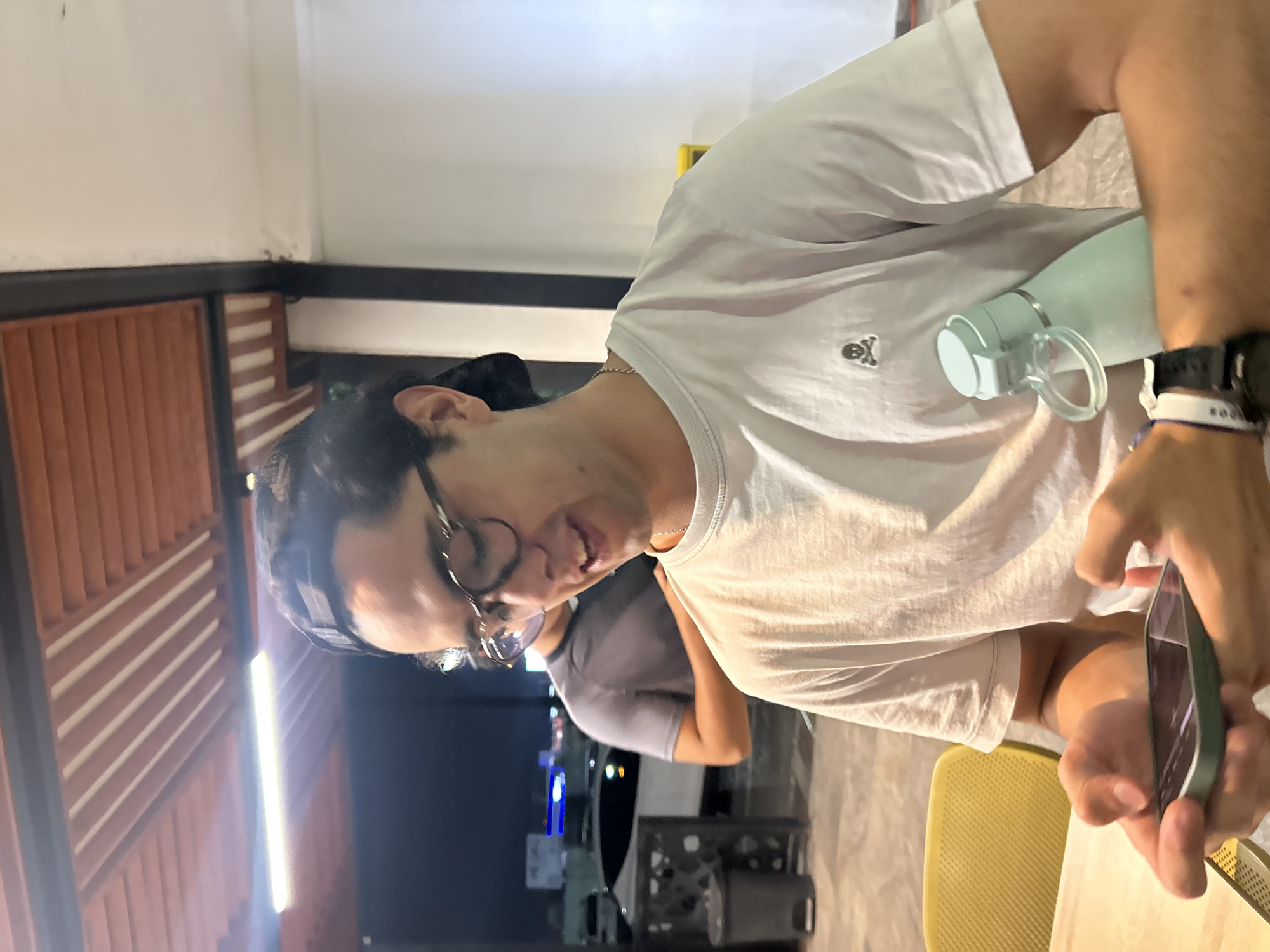 A photograph taken indoors shows a man seated at a table, engaged with his cell phone. The man has dark hair, glasses, and is wearing a white T-shirt with a small skull-and-crossbones graphic on the chest. He has a water bottle next to him. His wristwatch is visible. Another person can be seen in the background, wearing a gray shirt. The lighting is soft, and outside it is dark. The image provides no indications of the city.