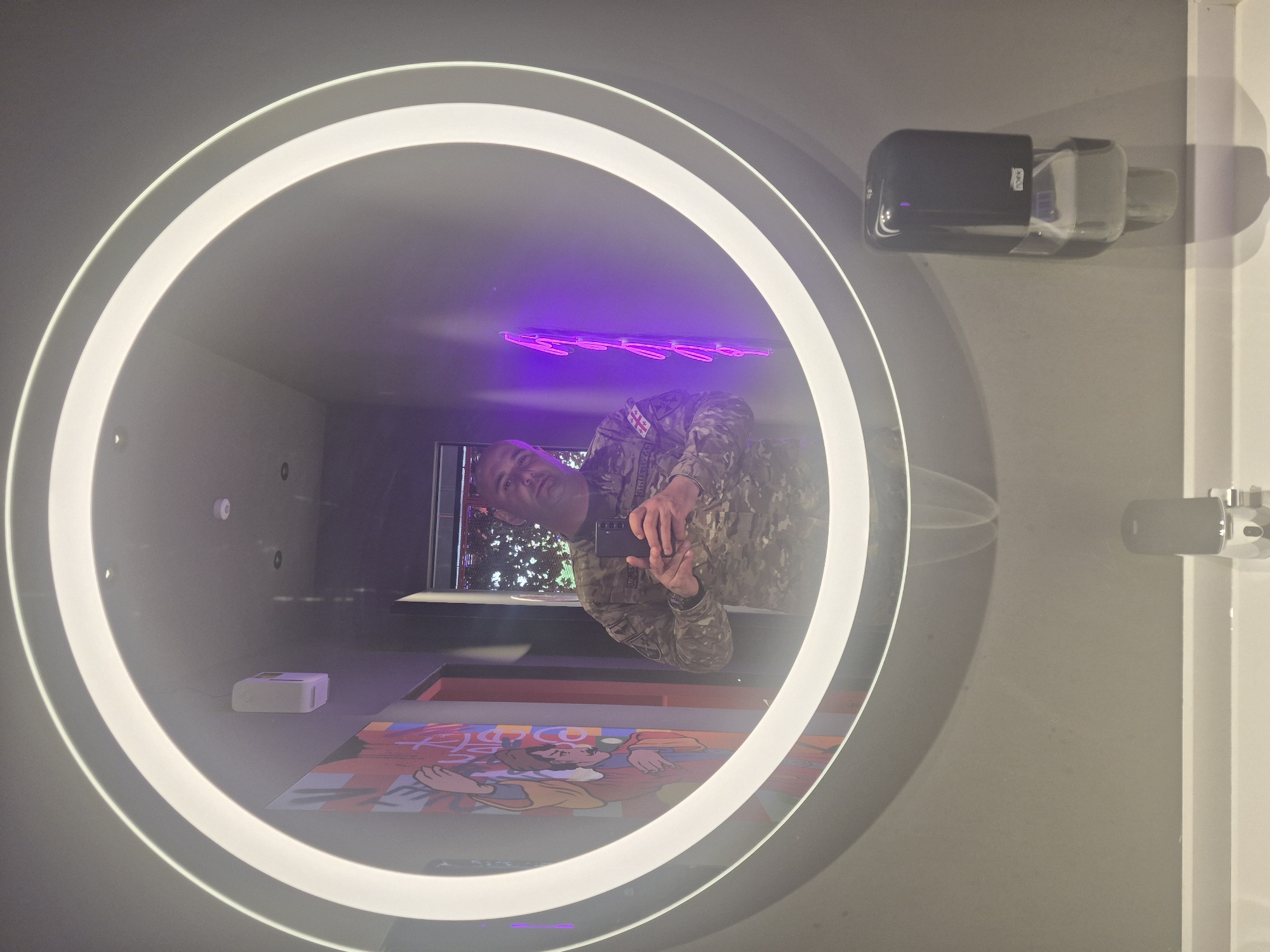 A mirror selfie shows a person reflected in a round mirror with a lit frame. The person, wearing a military uniform with Georgian flag patches, is holding a smartphone, presumably taking the photo. The mirror reflects a modern interior with a purple neon light fixture on the ceiling. Below the mirror is an image of an animated person lying on the floor. The background shows a window with trees visible. A soap dispenser and a faucet are mounted to the wall to the right of the mirror. Based on visual cues and a possible match, this is likely located in Fabrika Tbilisi, a multi-functional urban space in Tbilisi, Georgia.