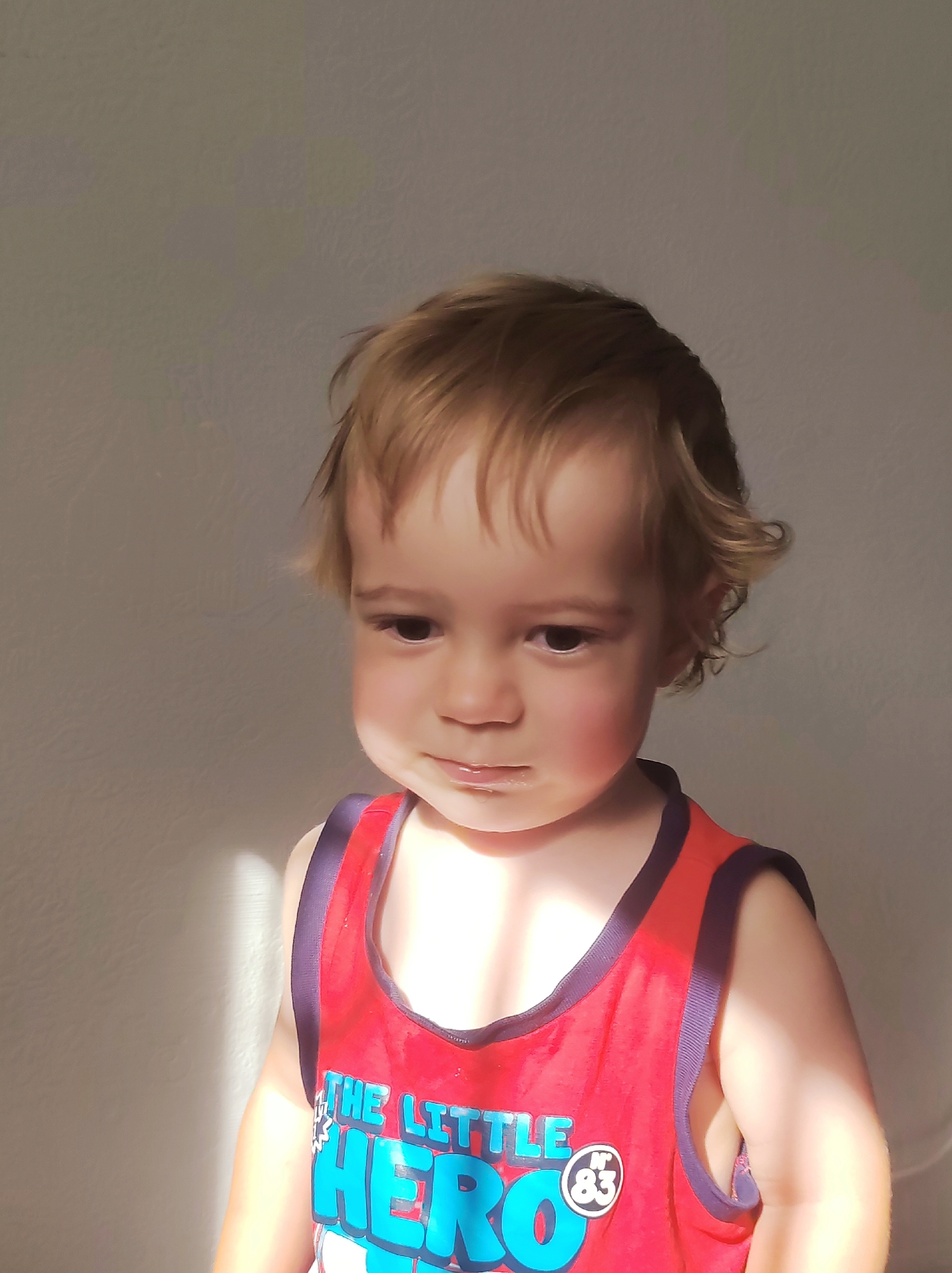 This is a waist-up, medium shot of a toddler boy facing slightly away from the camera, with his head turned to look downwards. The boy has fair skin, light brown hair styled in loose curls, and brown eyes. He is wearing a red tank top with navy blue trim. The front of the tank top has text that reads "THE LITTLE HERO" in large blue letters, with a circular emblem containing the number "83" to the right of the text. The background is a plain, textured wall that appears to be a neutral gray or off-white color. Sunlight casts a diagonal shadow across the left side of the wall and a patch of light on the boy's chest. The image appears to be taken indoors, possibly in a home environment. There is no discernible indication of the city of Chaikovsky, Russia, within the image content itself.
